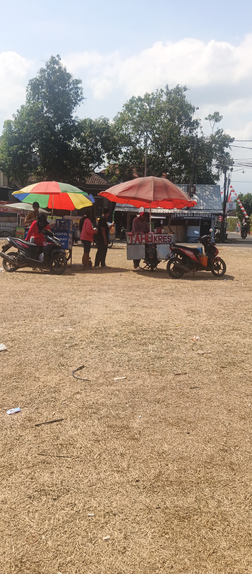 The image depicts an outdoor scene in what appears to be a public space in Gondang, Indonesia. Two food carts are set up, each under a large umbrella, one with a red umbrella and the other with a multi-colored umbrella (red, yellow, green). Several people are present, some interacting with the carts, likely as customers or vendors. A person in a red shirt is seated on a black motorcycle to the left of the multi-colored umbrella. To the right of the red umbrella, a red and black motorcycle is parked. The ground is covered in dry grass and scattered debris. In the background, buildings and trees are visible, along with some red and white checkered flags, suggesting a festive or event-related atmosphere. The sky is partly cloudy.
