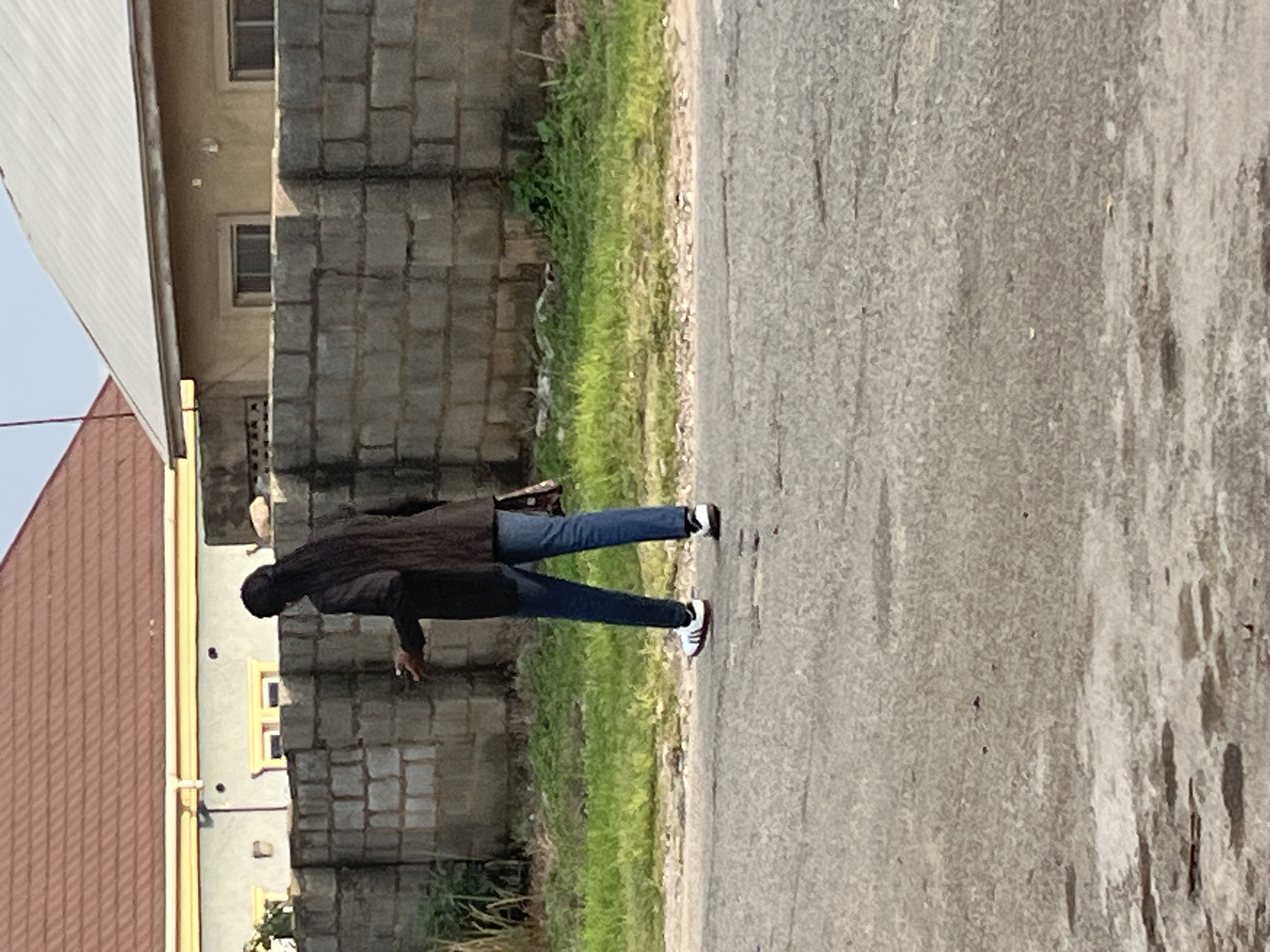 A person with long, dark hair is walking away from the camera on an asphalt road. They are wearing a dark jacket, blue jeans, and white athletic shoes with red accents. The person is carrying a dark bag over their right shoulder. Behind the person is a low-lying green grassy area with some vegetation. Beyond the grass is a wall constructed of light-colored concrete blocks. In the background, there are two residential buildings with tiled roofs, one with a red-tiled roof and the other with a grey, corrugated metal roof. The buildings have windows and are painted in neutral colors. The scene appears to be in a residential area of Abuja, Nigeria, characterized by the architecture and materials used for the buildings and wall.