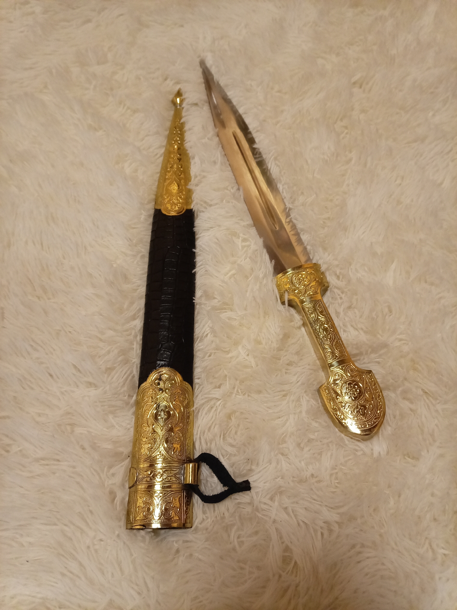 This image displays a dagger with a golden, ornate handle and a sheath of black material, also adorned with golden embellishments. The dagger and sheath are laid out on a white, fluffy surface. The scene is indoors, with no discernable urban or environmental features visible that could indicate a specific location in Yessentuki, Russia. There are no people or actions depicted.