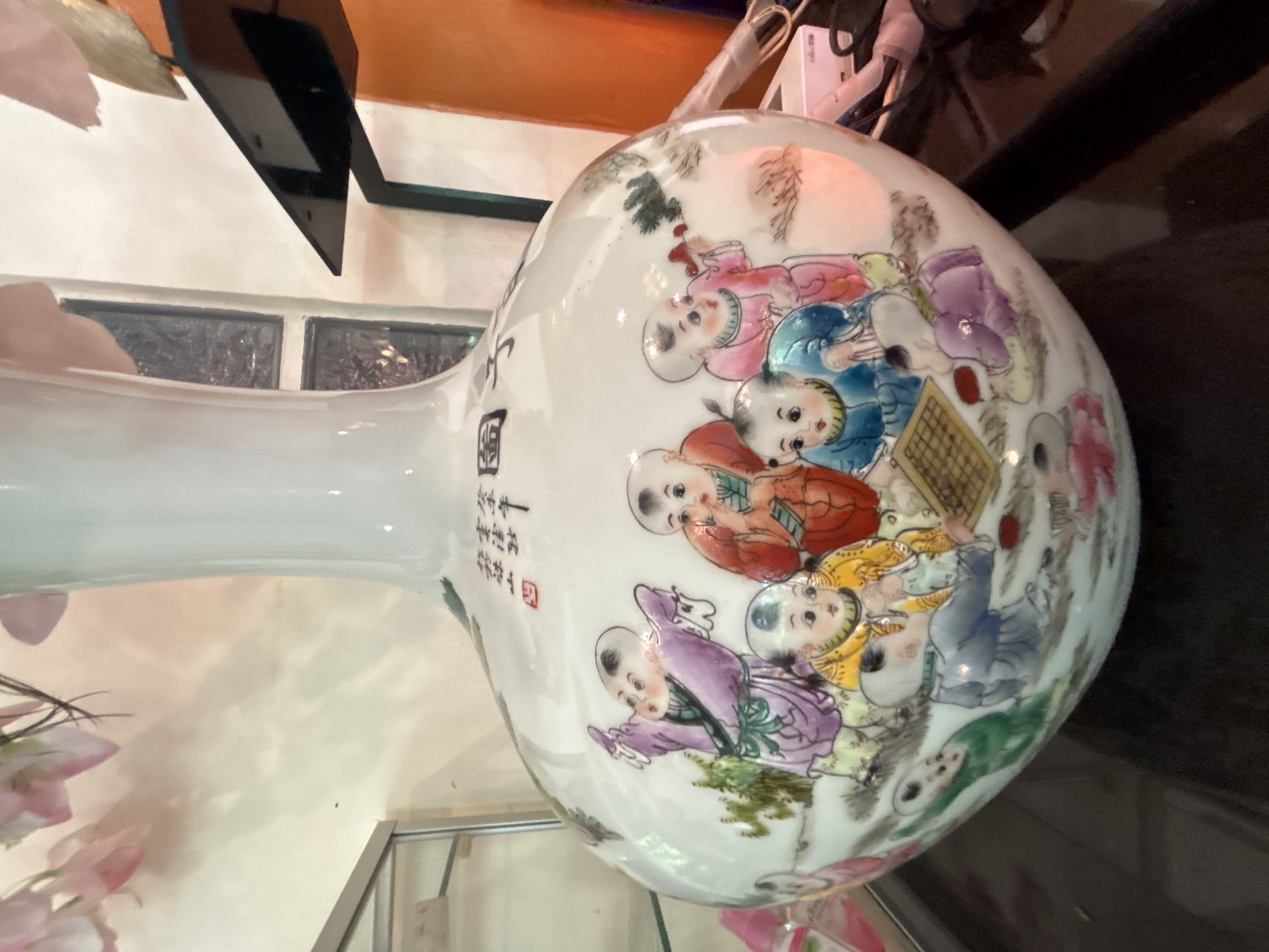 The image displays a white ceramic vase adorned with a vibrant painting of children playing a board game. The children are depicted in various colorful garments and engage in the game with concentration. Chinese calligraphy is visible at the top of the vase, near its neck. The vase is placed on a dark surface, possibly a table, and is partially illuminated by soft lighting. In the background, a shelf with various items and a window with a textured glass pane can be observed. The scene appears to be indoors, likely in a home or shop environment. The specific location in the city of Chalong, Thailand, is not discernible from the visual information provided in the image.