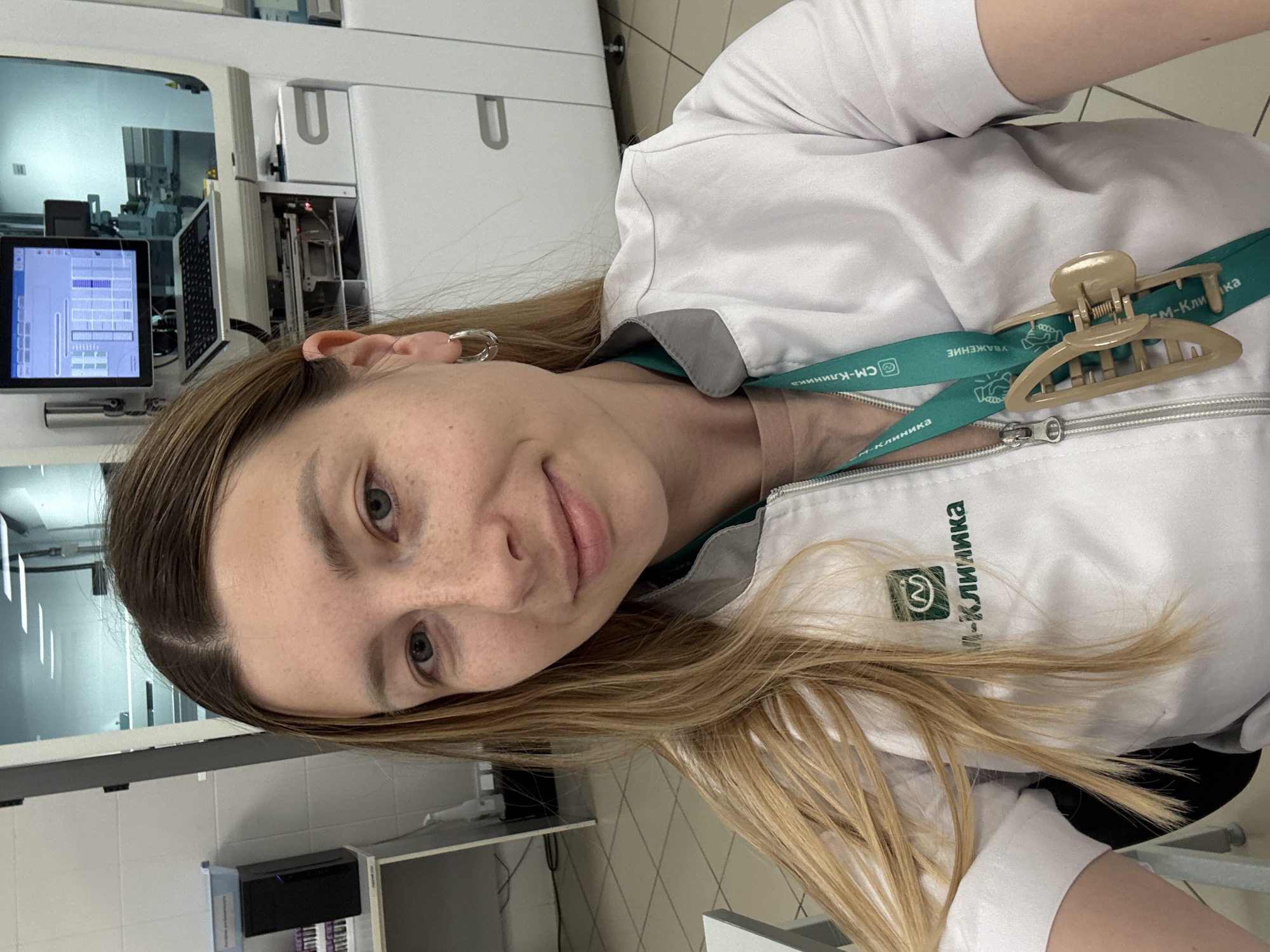 A woman wearing a white medical coat and a green lanyard with Russian text stands in what appears to be a medical laboratory. The text on the coat reads "СМ-Клиника," suggesting she works for the SM-Clinic medical network. In the background, there is medical equipment, including a machine with a screen displaying graphical data. The environment looks sterile, with tiled walls and modern medical apparatus. Considering the text on her uniform, the location is likely a branch of the SM-Clinic in Moscow, Russia.