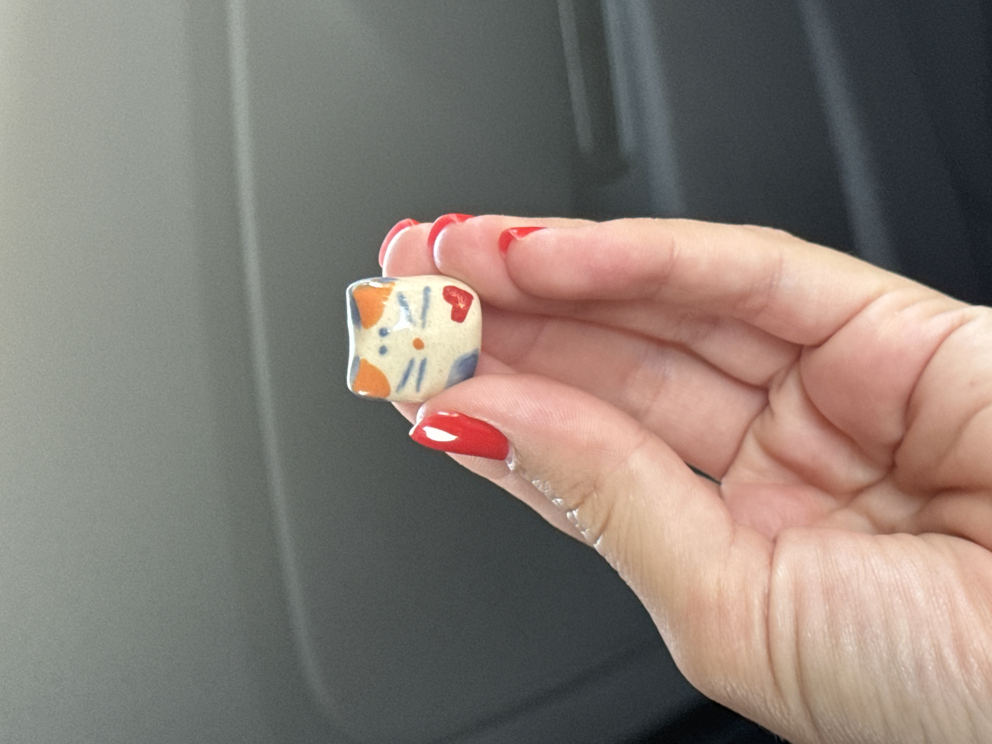 A hand with bright red fingernail polish holds a small, ceramic cat figurine. The figurine is white with orange ears, blue markings on its face, and a small red heart on its chest. The background is dark and out of focus, suggesting an indoor setting or a car interior. The image does not provide enough information to determine a specific location within Podgorica, Montenegro.