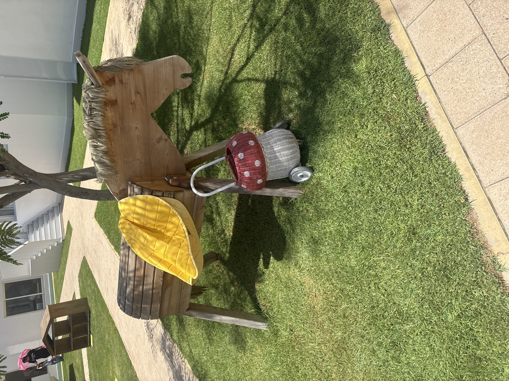 A wooden rocking horse sits on a patch of vibrant green grass, partially shaded by a nearby tree. The horse has a segmented wooden body and a mane made of light-colored yarn. A bright yellow, quilted cushion is draped over the horse's back. To the right of the horse, a small, woven basket shaped like a mushroom with a red and white polka dot top and white base sits on wheels. In the background, a paved walkway curves around the grassy area. Further in the background, a modern building with white walls and large windows is visible, along with a wooden playhouse and a person holding a pink umbrella. The scene appears to be an outdoor play area, possibly part of a school or a residential complex, in Dubai, United Arab Emirates.