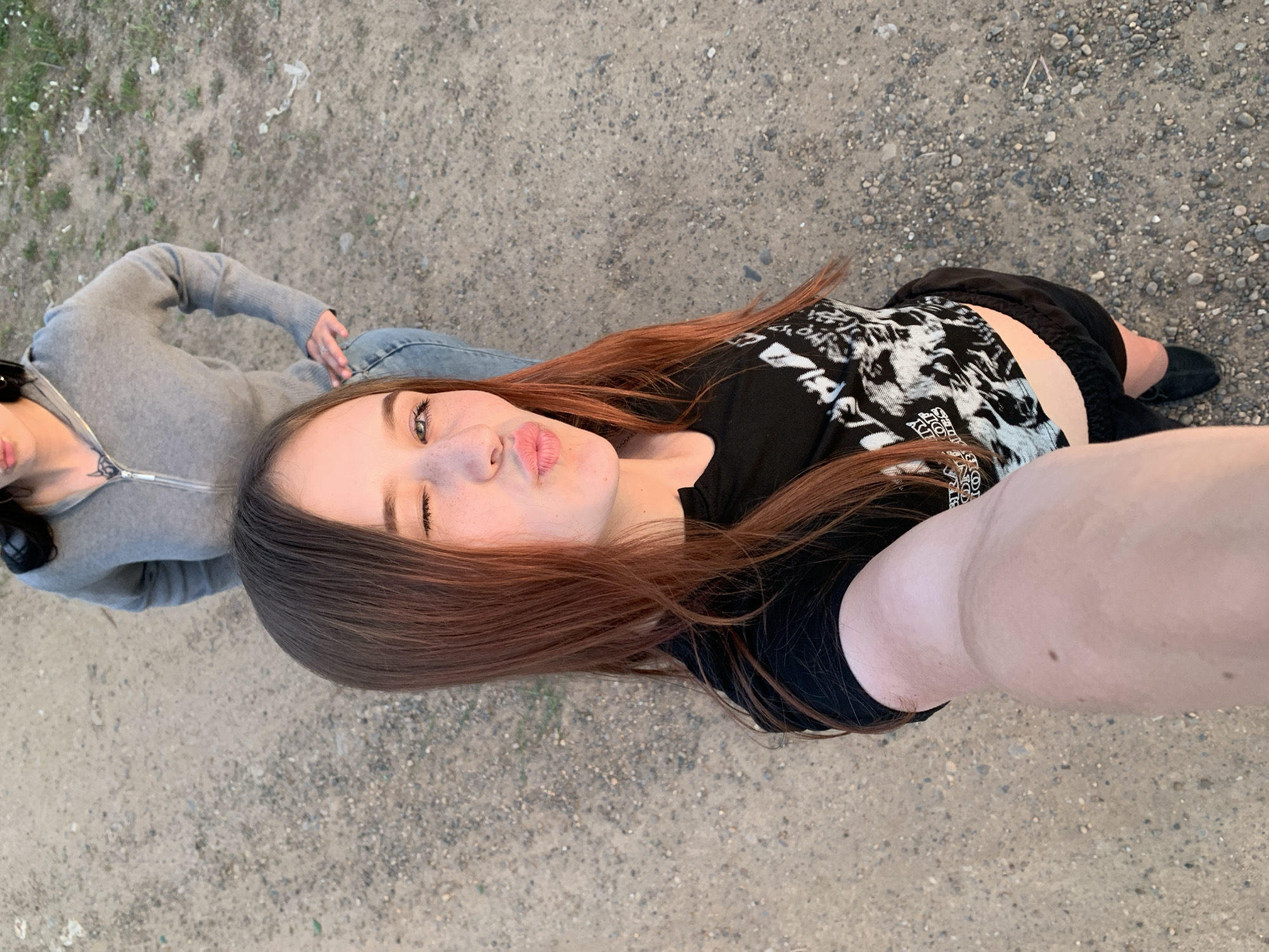 Two people are on a gravel surface. The person closest to the camera is a young person with long red-brown hair, wearing a black shirt and black shorts, taking a selfie with their arm extended. They are looking at the camera with pursed lips. The other person is wearing a gray hooded sweatshirt and jeans and is standing slightly behind and to the left of the first person. The ground is gravel. The location is outdoors in a city like Фоки, Russia.