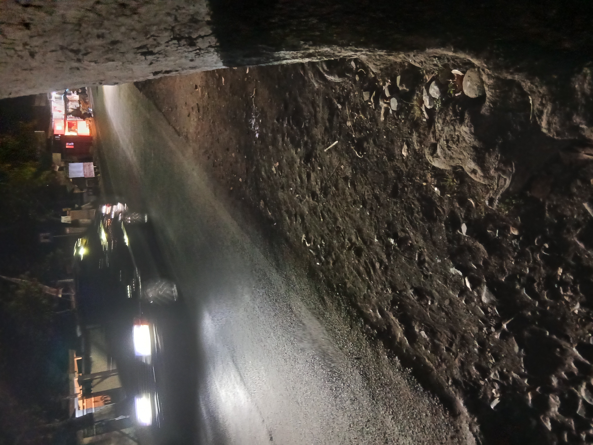 The image depicts a nighttime scene on a road in Kademangan Kulon, Indonesia. A dark-colored car with its headlights on is moving on the asphalt road. The road surface appears wet, reflecting the car's headlights. To the right of the road, a large tree trunk is prominently featured, its rough bark visible. The ground beside the road is unpaved and covered with dirt, rocks, and some scattered leaves. In the background, across the road, there are several small buildings or stalls, one of which is illuminated with bright red and orange lights, suggesting it might be a food stall or shop. The overall lighting suggests it is nighttime, with artificial lights from the car and the stalls illuminating the scene. The environment appears to be a rural or semi-urban area, given the presence of the unpaved ground, tree, and smaller structures.