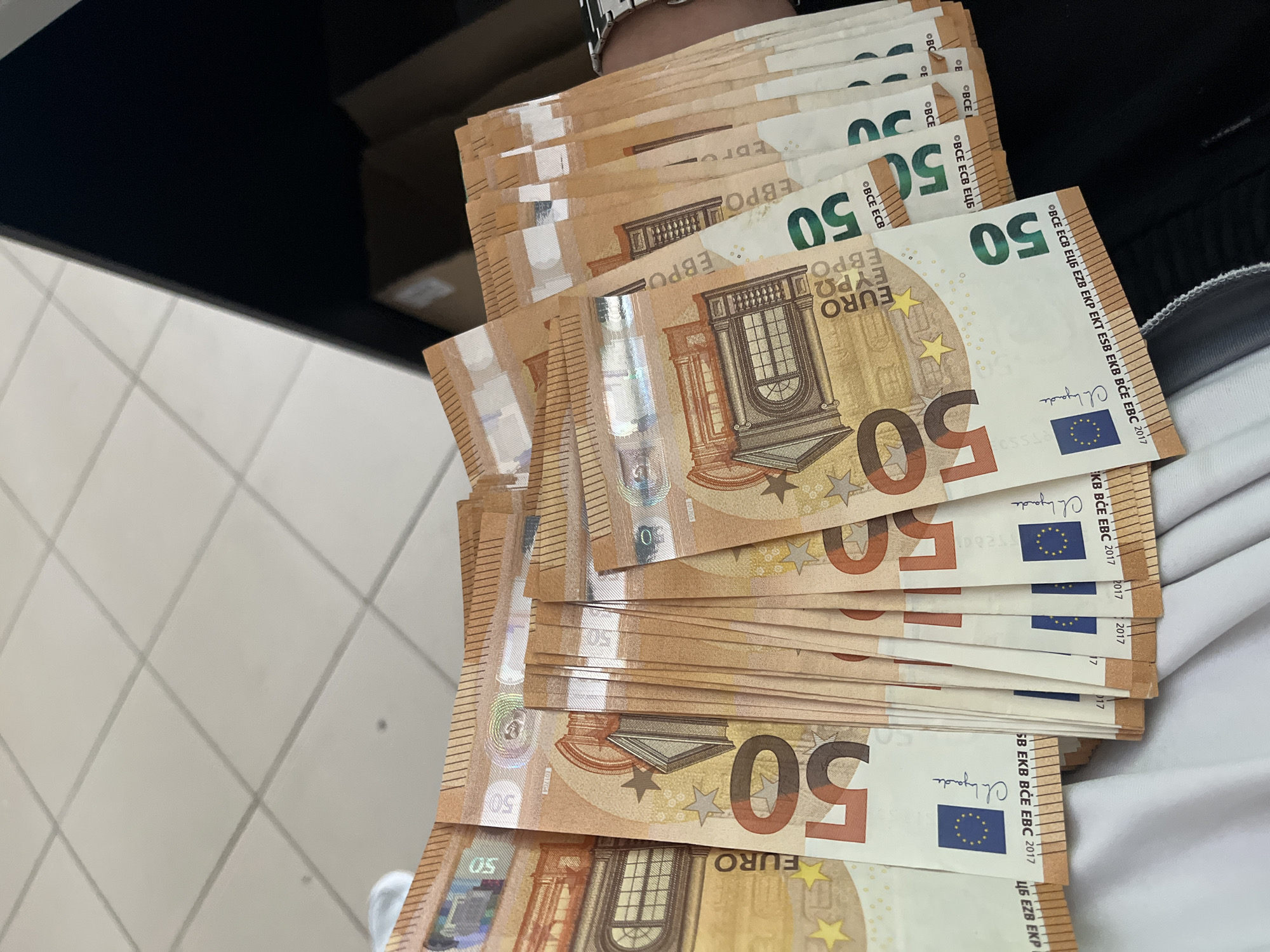 A high-angle, close-up shot captures a fanned-out stack of numerous 50 Euro banknotes held by a person. The notes are primarily orange and brown, displaying architectural elements, the European Union flag, and a hologram featuring the number "50" and iridescent patterns. Most visible notes are crisp and appear new.
Partial views of a person's wrist, adorned with a metallic silver-colored watch or bracelet, and a white sleeve or piece of clothing, are visible in the upper and right portions of the frame, indicating they are holding the currency.
The background consists of a dark, possibly black, surface in the upper left, partially obscured by the notes. Below and to the left, a light-colored tiled floor with gray grout lines is visible. A blurred brown cardboard box can be faintly seen in the distance in the upper background.
The activity depicted is the holding or displaying of a significant quantity of 50 Euro notes.
Visible text on the banknotes includes "50", "EURO", "ΕΥΡΩ", "EBPO", "ЕЦБ", "BCE ECB", the full sequence "BCE ECB EZB EKT ESB EKB BCE EBC 2017", and "2017". The signature "Ch. Lagarde" (Christine Lagarde, President of the European Central Bank) is also discernible, along with partial serial numbers like "017G4" or "01764". The location context of Ivailo, Bulgaria, suggests these are local currency notes.