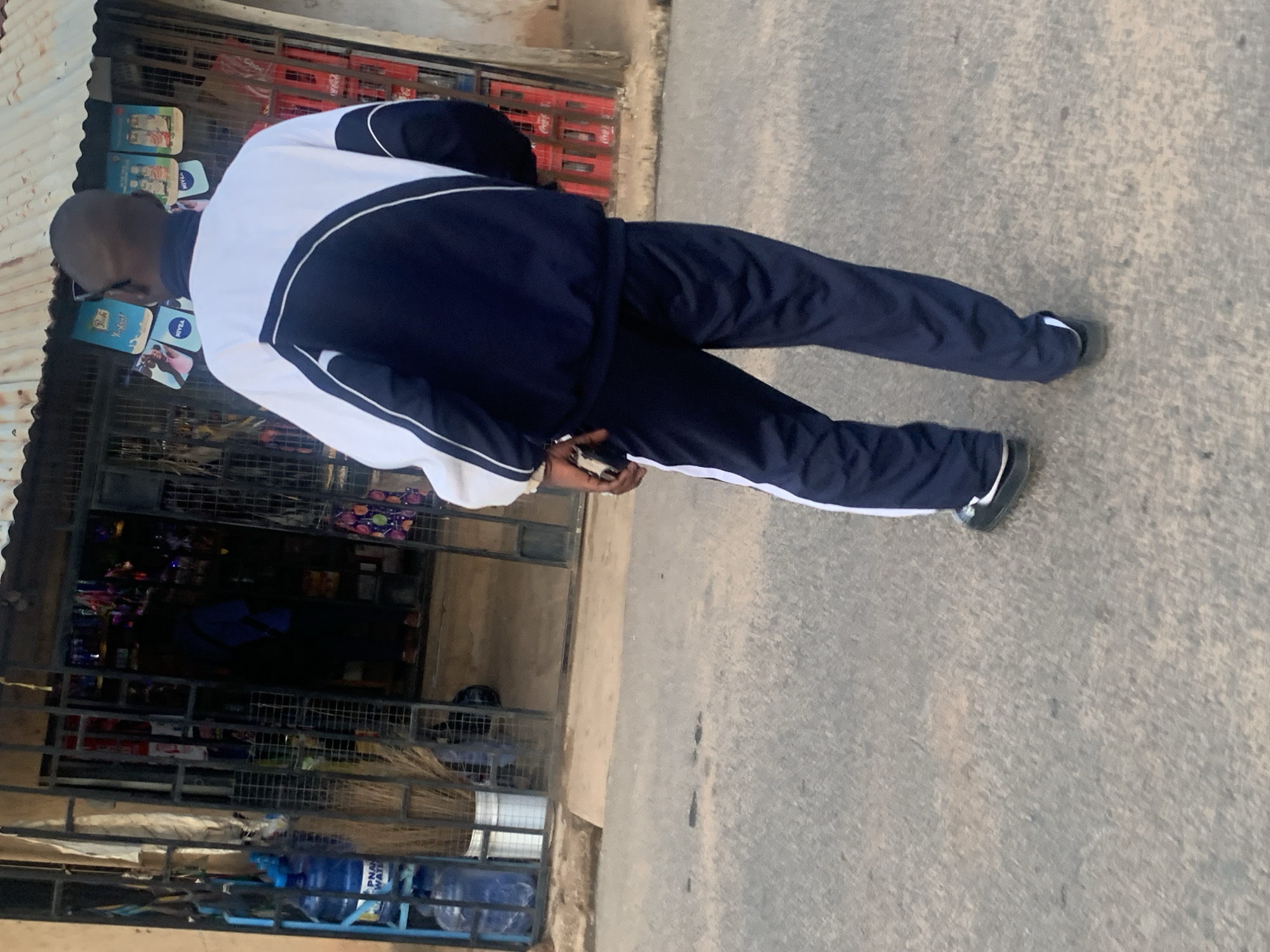 An outdoor, medium-shot image captures a person standing on a paved street next to a roadside shop in what appears to be Barakin Akawo, Nigeria. The individual, seen from behind and slightly to the side, is wearing a dark blue tracksuit with white panels and stripes, and dark shoes with white soles. They hold a dark object, possibly a mobile phone, in their left hand.
The setting is a bustling daytime street scene, with a small shop or kiosk to the left. The shop features a corrugated metal awning and is secured by metal grates. Various products are visible both inside and displayed on the exterior. On an upper shelf outside, multiple blue and white Nivea cosmetic products and a package possibly branded "Pears" or "Peal's" are arranged. Inside the shop, behind the grates, shelves are stocked with bottled drinks, including red Coca-Cola crates, and other packaged goods. Several large blue containers of "PNAP WATER" are visible towards the bottom of the shop's display area, along with brooms. The ground is a light gray, textured paved surface. The lighting suggests it is daytime with clear weather.