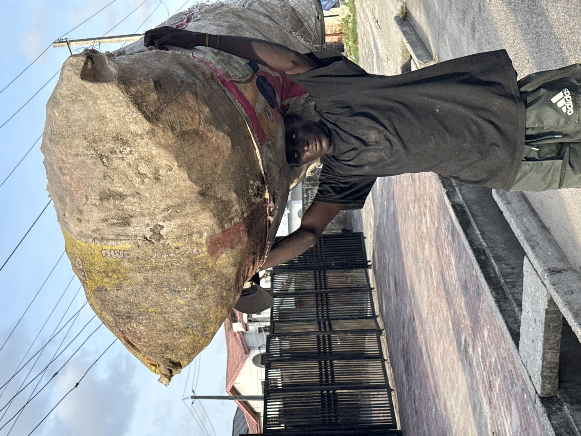 In Ado, Nigeria, a man in a black t-shirt and Adidas athletic pants carries a large, dirty sack on his back. He is positioned on a paved road adjacent to a building with a gated, barred entrance. Overhead, power lines are visible against the sky.