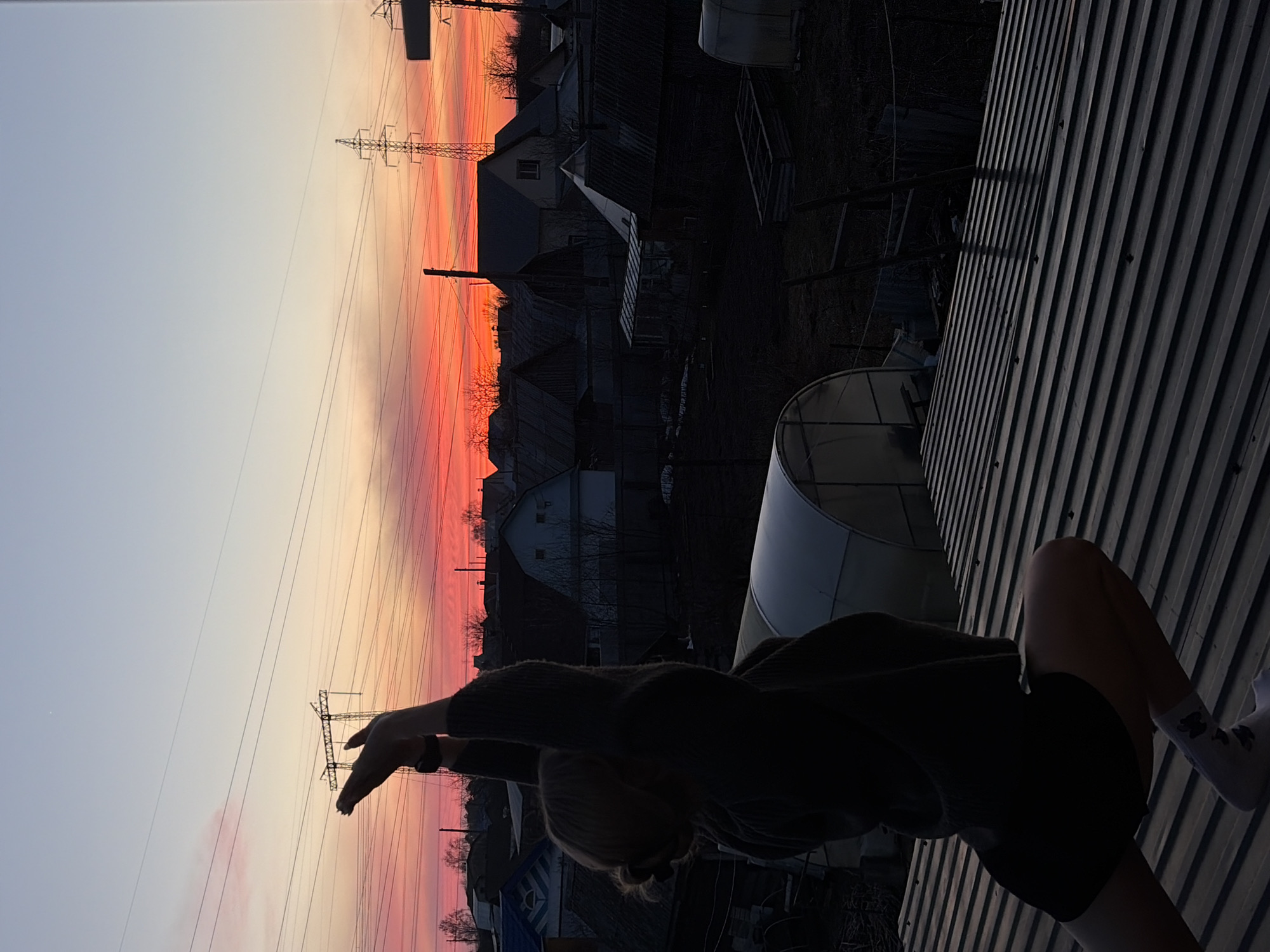 A medium-angle, eye-level shot, rotated 90 degrees counterclockwise, depicts a person sitting on a corrugated metal surface under a vibrant sunset sky in Chaikovsky, Russia. The sunset exhibits hues of orange, pink, and yellow, with a gradient transitioning into a light blue at the top. A silhouette of electrical pylons and power lines is visible against the bright sky, along with silhouetted buildings and bare tree branches.
The person, also silhouetted, is seated on the corrugated metal, with their back to the viewer, wearing a dark top, shorts, and socks. Their arms are raised towards the sky, and the person is likely engaging in some sort of stretching or reaching motion.
The corrugated metal surface occupies the lower right quadrant of the frame and obscures some of the view.
The general location is identified as Chaikovsky, Russia, based on analysis of the architecture and visible infrastructure.