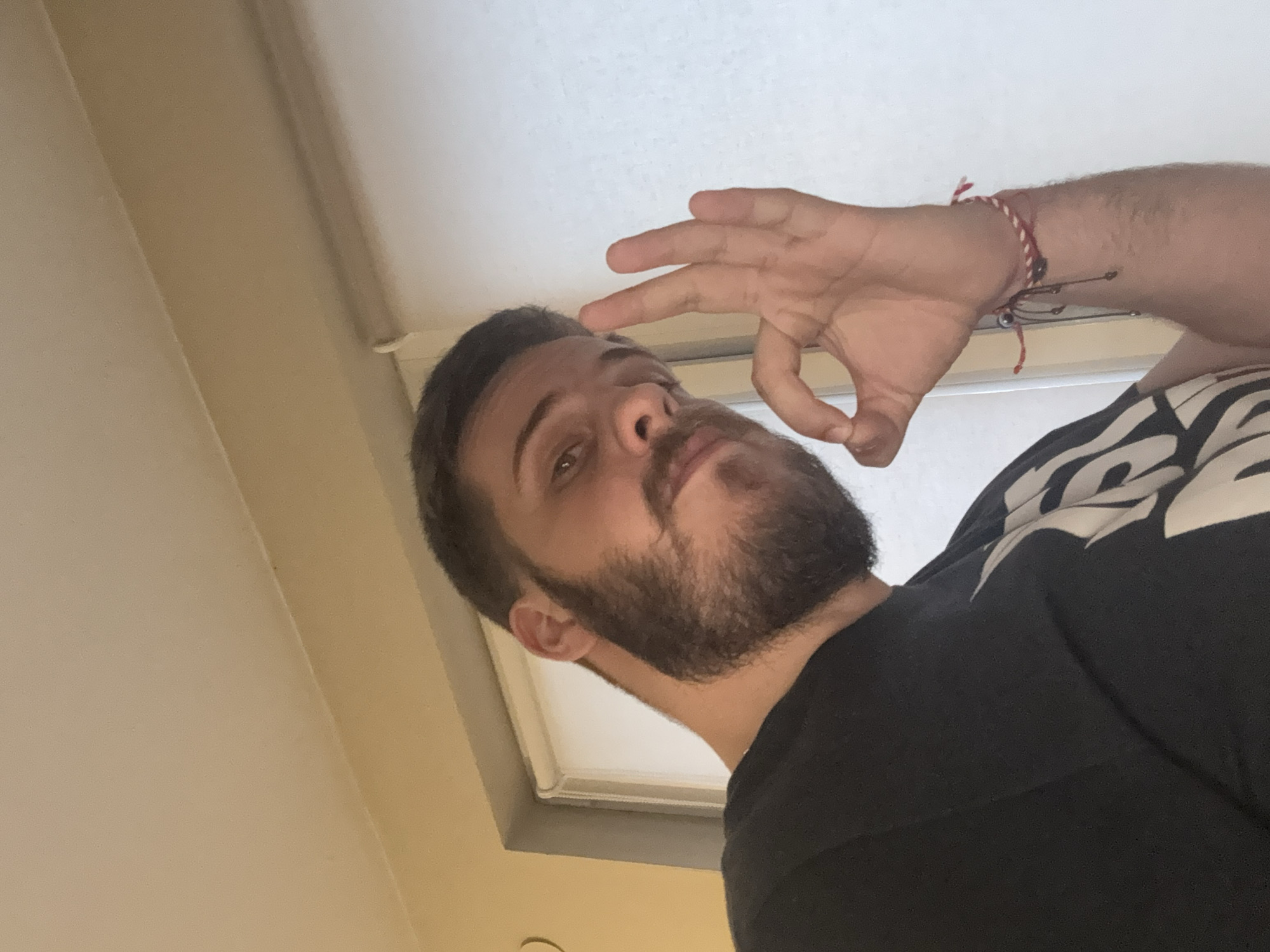 A bearded man, presumably indoors, is making an "OK" hand gesture. He is wearing a black t-shirt. A wrist bracelet of red and white string and another string bracelet with attachments is on his left wrist. The background shows parts of a white or off-white wall or ceiling and a window frame. The specific location within Sofia, Bulgaria cannot be determined from the visual information provided in the image.