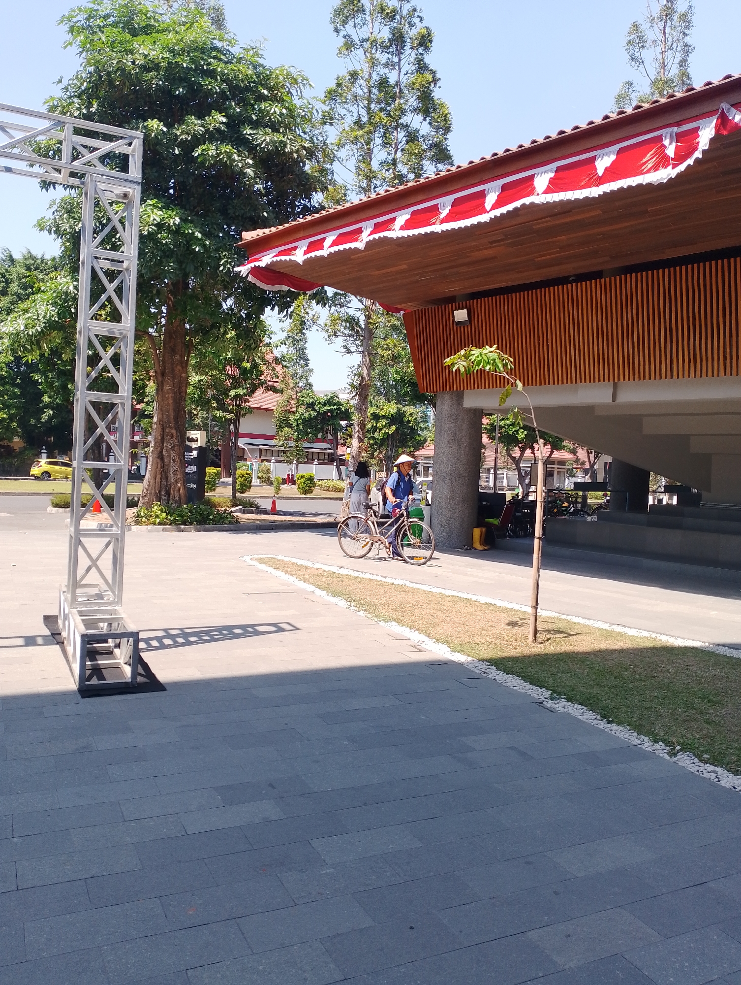 The image shows an outdoor scene in Depok, Indonesia, likely a public area near a building with an overhang. In the foreground, a metal truss structure is visible on the left, casting a shadow. The ground is paved with large, dark gray rectangular tiles. To the right, there is a patch of grass bordered by white pebbles and then more paving.
In the mid-ground, a person wearing a conical hat and a blue shirt is standing next to a bicycle. The bicycle appears to be vintage-style and is brown. Another person is partially visible behind the cyclist, also with a bicycle. Behind them, a road is visible with a yellow car driving on it and a red traffic cone.
Further back, a large building with a wooden facade and a red and white draped awning is prominent. Trees with lush green foliage are scattered throughout the scene. The sky is clear and blue, indicating a sunny day. The overall impression is that of a well-maintained urban outdoor space.