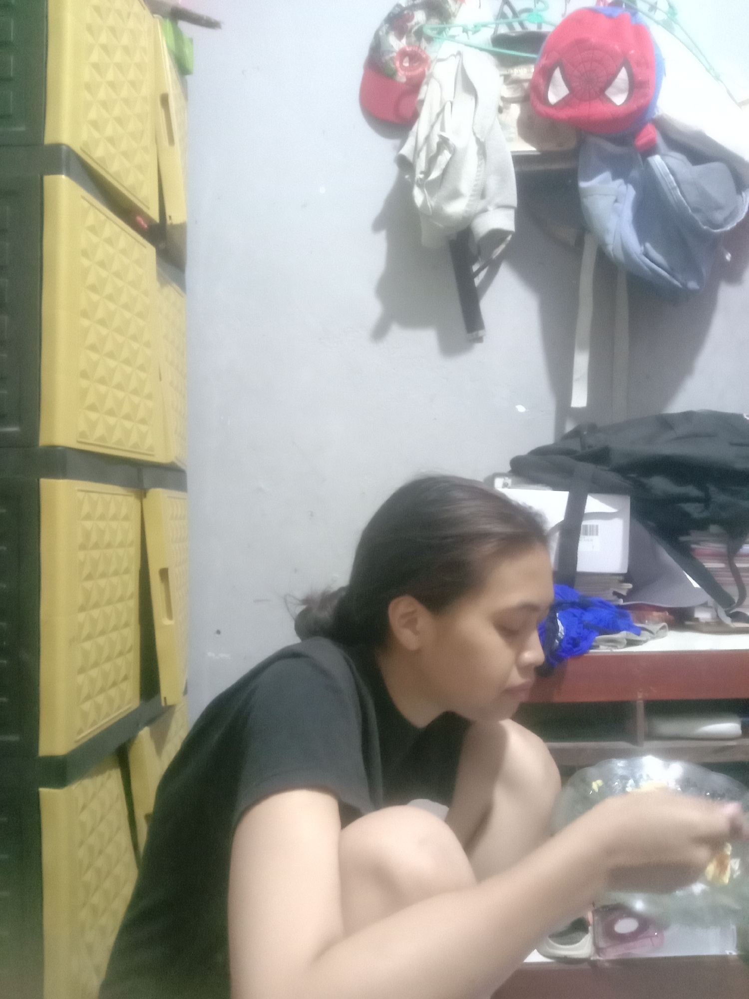 The image depicts a young woman with dark hair, wearing a black t-shirt, squatting in a room. She is looking down at a clear bowl held in her hands, which appears to contain food. To her left is a stack of yellow plastic drawers. In the background, a wall is visible, with various items hanging or placed on shelves, including a red Spider-Man plush toy and a grey hoodie. The lighting in the room is dim. The scene suggests an indoor domestic setting.