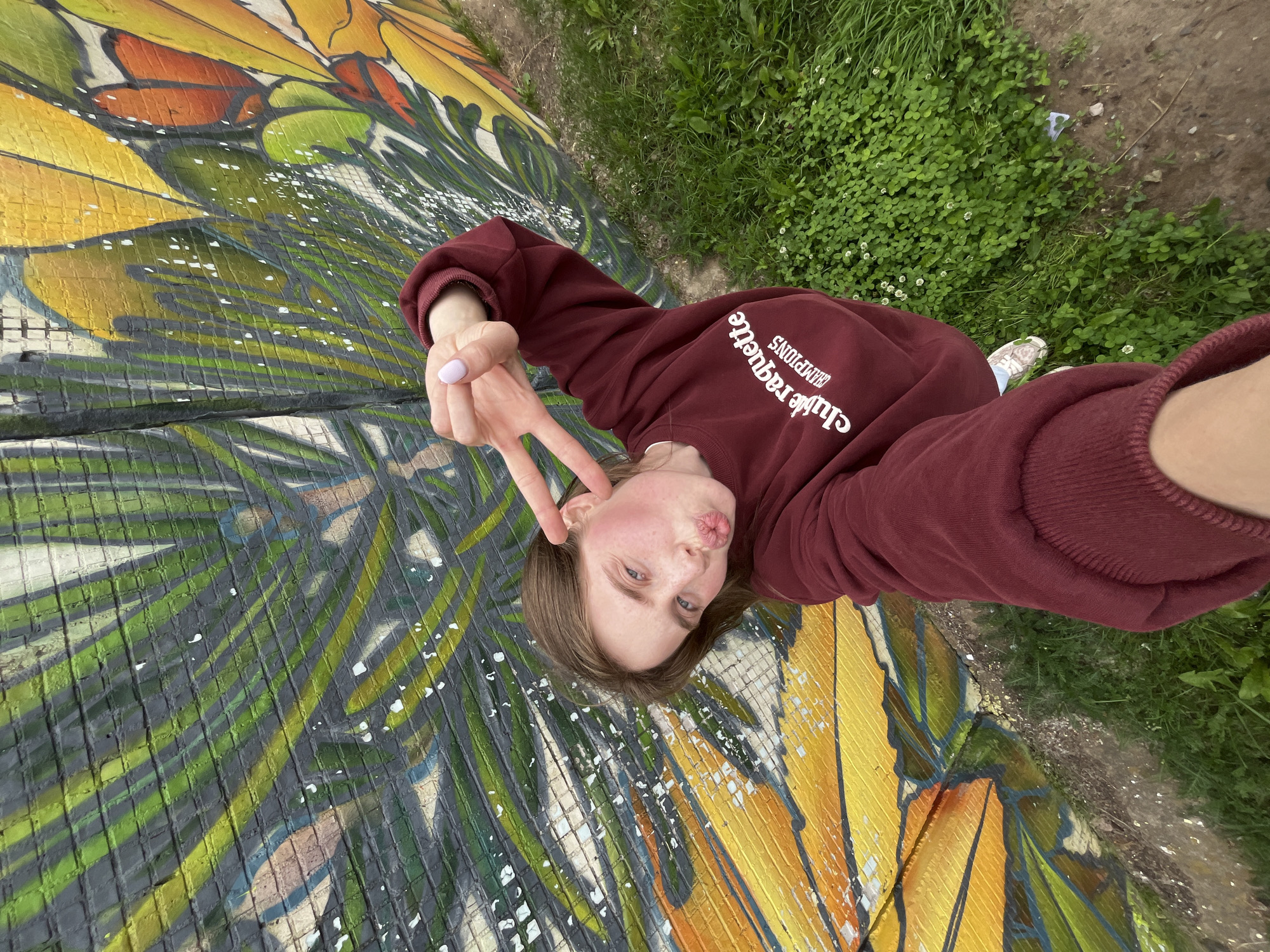 A young woman is lying down and taking a selfie on a colorful mural that resembles a butterfly wing. The mural features vibrant shades of yellow, orange, green, and red, with a textured, grid-like pattern overlaying it. The woman is wearing a maroon sweatshirt with white text on the front that reads "club raquette CHAMPIONS". She is making a peace sign with her right hand and puckering her lips for the camera. Her left arm is extended, holding the phone. The setting appears to be outdoors, with green grass and dirt visible in the background. The lighting suggests it is daytime, and the weather seems pleasant. The overall mood is playful and relaxed. The location is identified as Chaikovsky, Russia.