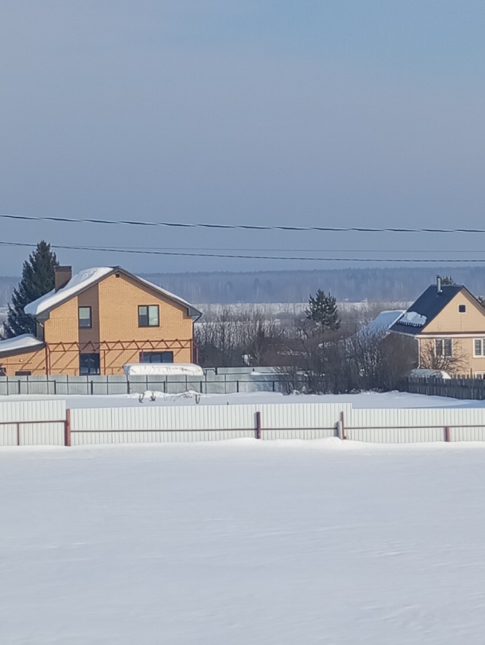 This image depicts a winter residential landscape in Яромаска, Russia, under a clear, pale blue sky. The foreground consists of a vast, unbroken expanse of white snow. In the midground, a white corrugated metal fence with dark brown posts runs horizontally, partially submerged in the snow.
Beyond the fence, two houses are visible. On the left, a two-story residence with light brown brick walls and a dark, snow-covered gabled roof stands. It features multiple dark-framed windows, including one on the upper floor and several on the lower. A dark green coniferous tree is situated to the left of this house. Behind its immediate property fence, a white, tarp-covered object, possibly a boat or vehicle, is discernible on the snow.
To the right, another house is partially visible, also featuring light-colored walls and a dark, snow-dusted gabled roof. A line of bare trees and shrubs separates the houses from the immediate foreground. In the distant background, a faint, dark line indicates a forest or land horizon, meeting the clear sky. Two dark power lines extend horizontally across the upper portion of the image. No individuals are present in the scene.