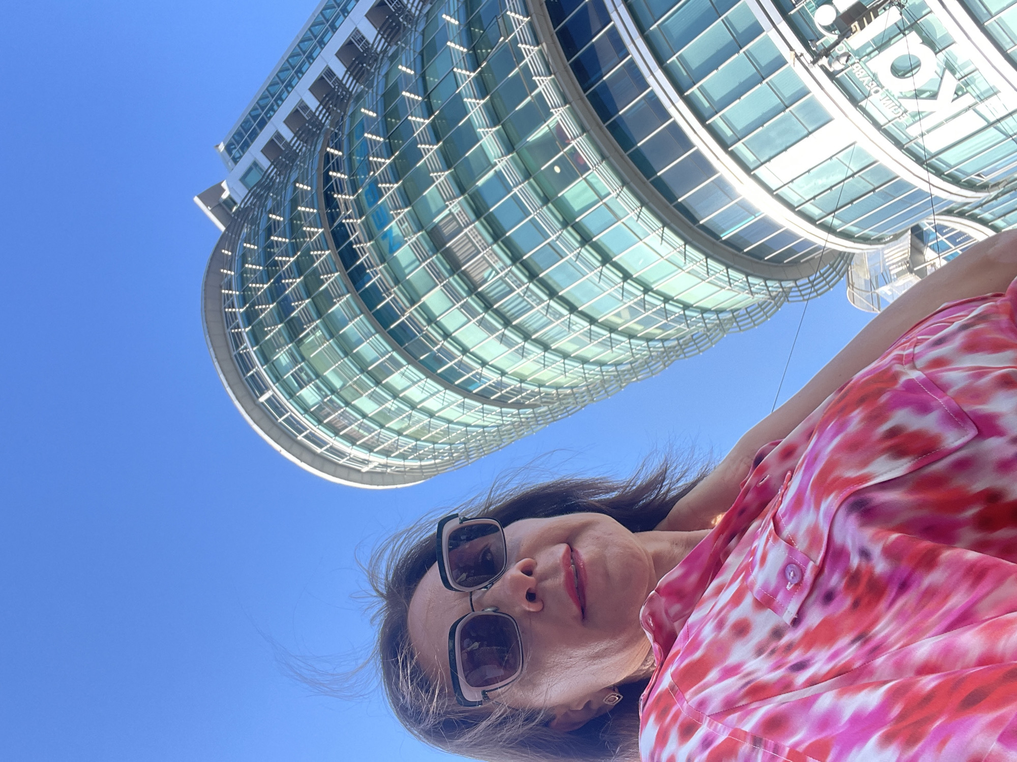 A low-angle, upward-looking selfie shows a woman with brown hair and sunglasses posing in front of a modern, curved glass building under a clear blue sky. The woman is wearing a pink and red tie-dye patterned shirt. The building features a reflective, curved glass facade with horizontal silver accents. The sun is shining brightly, casting shadows on the building. The time of day appears to be daytime, likely mid-morning or afternoon, given the bright sunlight. The scene suggests an urban setting, possibly a business or entertainment district. The woman's expression is neutral, and the overall mood is casual and outdoorsy. The building has text visible on its upper section, which appears to be illuminated white letters. The text reads "KOLOSOV", "V.I. LENIN", and "EQUALLY OBVIOUSLY". The image is likely taken in Sochi, Russia, based on the provided location context.