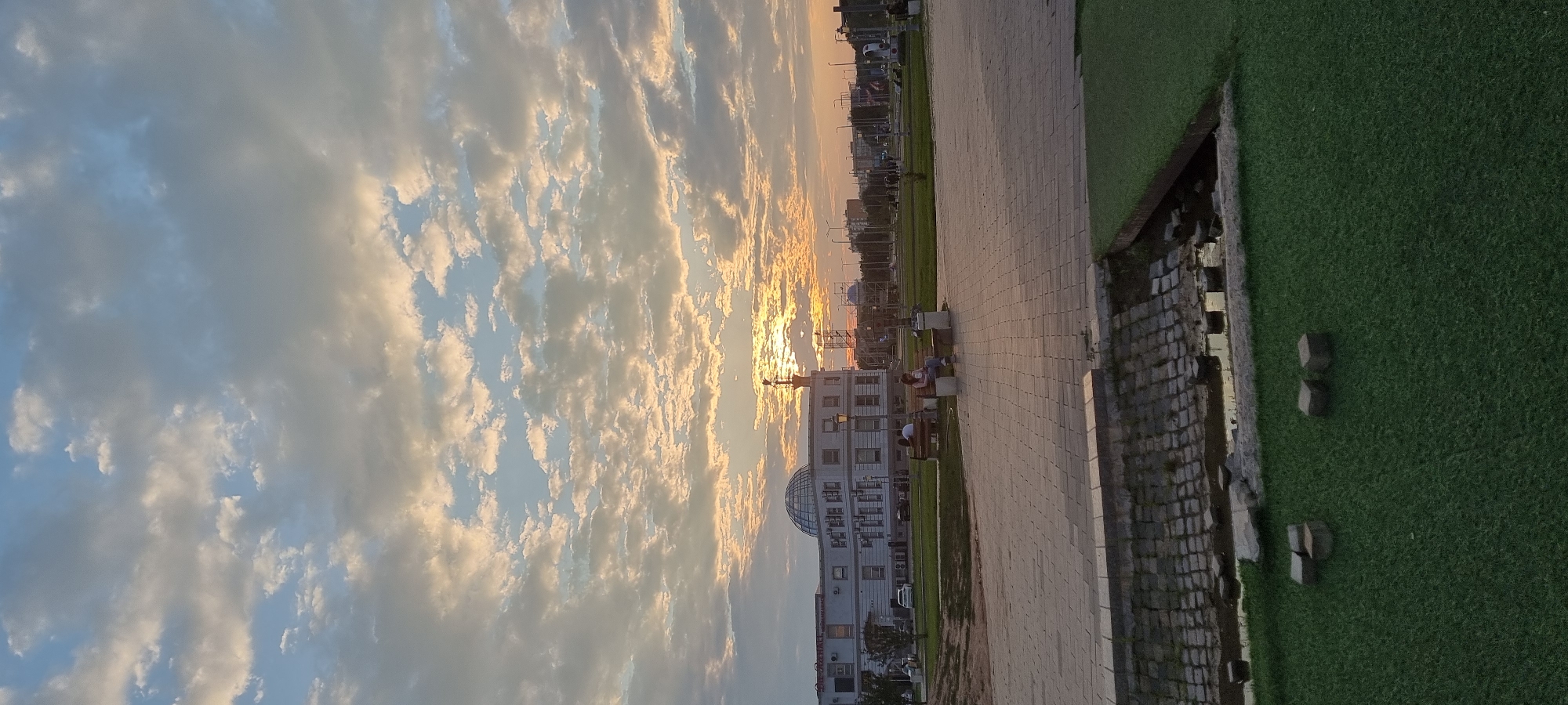 The image captures a serene sunset scene in Aktobe, Kazakhstan. The sky is filled with a dramatic display of clouds, tinged with the warm hues of the setting sun. Streaks of orange and yellow intermingle with shades of blue and white, creating a painterly backdrop.
In the foreground, a paved walkway runs parallel to a lush green lawn. The walkway is bordered by a stone wall and a small body of water. Scattered along the walkway are several benches, two of which are occupied by people. One bench has two individuals sitting, appearing to be engaged in conversation. Another bench is occupied by a single person.
Beyond the walkway and lawn, a modern white building with a distinctive dome-shaped structure stands prominently. The building's architecture suggests it might be a public or commercial establishment. The setting appears to be an urban park or a public space designed for leisure and relaxation, given the presence of the walkway, benches, and green areas.
The overall mood is peaceful and contemplative, enhanced by the soft, warm light of the sunset. The time of day is clearly evening, as indicated by the sun's low position and the colors in the sky. The weather appears clear and pleasant, conducive to people enjoying the outdoors. No visible text is discernible in the image.