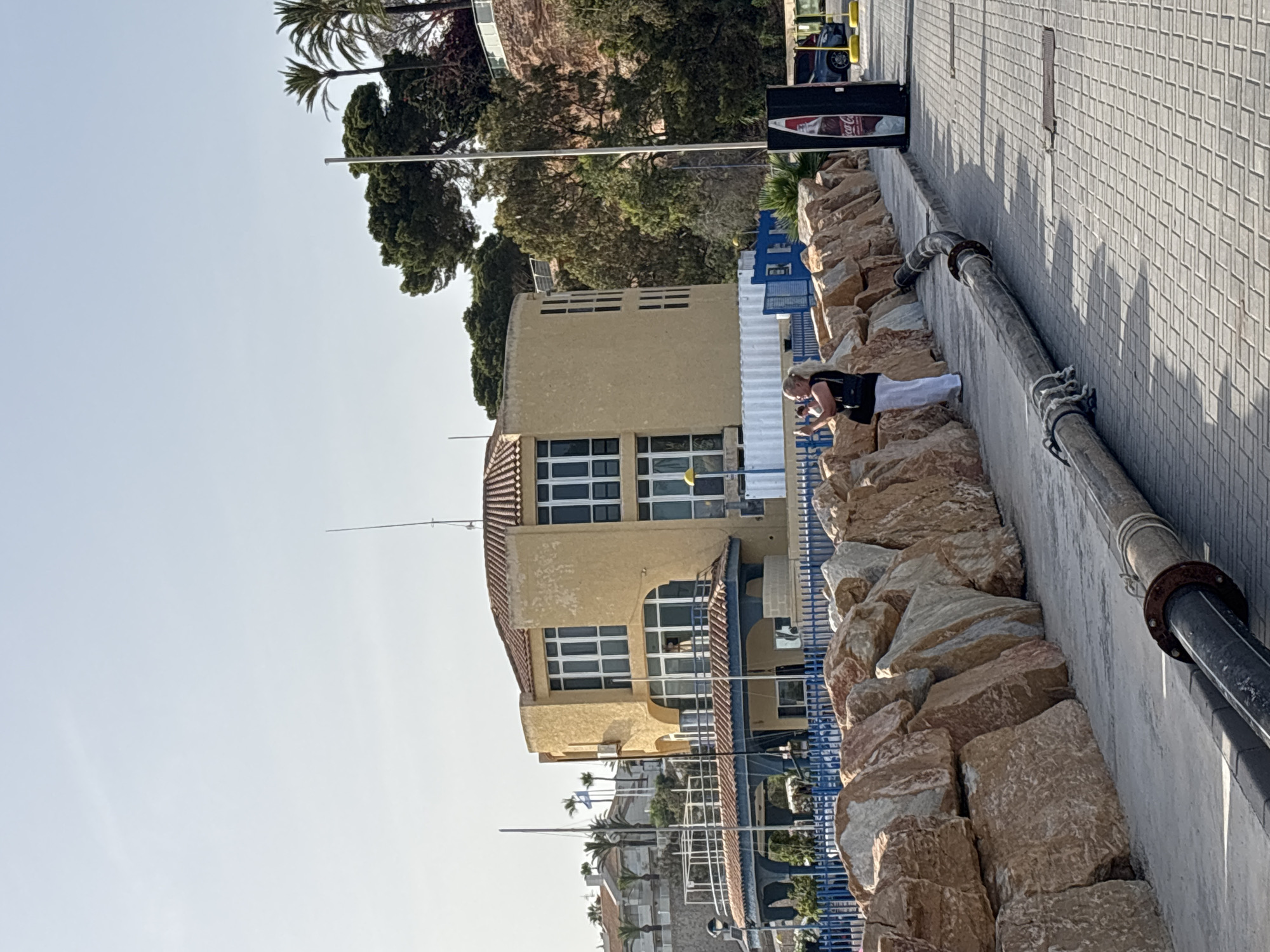 A woman stands on a paved walkway next to a wall of large, rough-hewn rocks. She is wearing white pants and a black top, and is holding a phone up, appearing to take a picture. Behind her, a multi-story, ochre-colored building with a red-tiled roof features large windows and arched doorways. A blue fence separates the walkway from the building's grounds. In the background, palm trees and other vegetation are visible on a hillside. A large, flexible black pipe runs along the edge of the walkway, secured by ropes at intervals. The scene appears to be a coastal area in Urbanización Cabo Roig, Spain, under a clear blue sky.