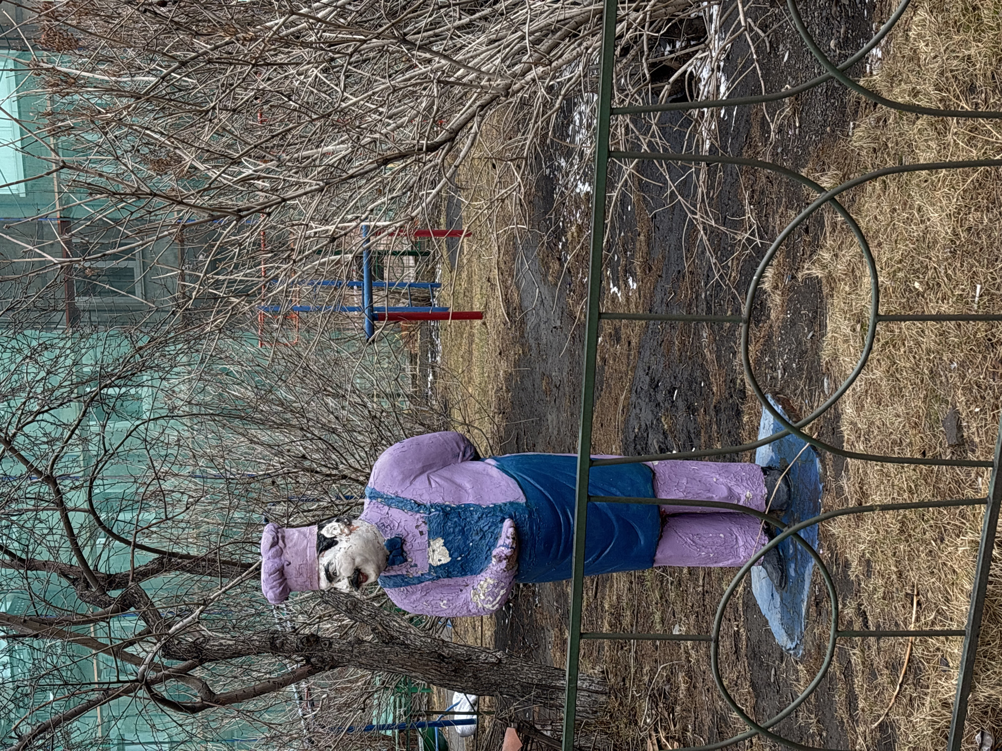 The image depicts a weathered, painted statue of a cook or chef in a city park or similar location, likely in Unknown. The statue is primarily purple with blue overalls and shoes. It stands on what appears to be a lawn of dormant grass and bare earth, with bare trees surrounding it. Part of a green metal fence is visible in the foreground. A building can be seen through the trees in the background, along with some playground equipment. The statue appears aged, with paint peeling off in places.