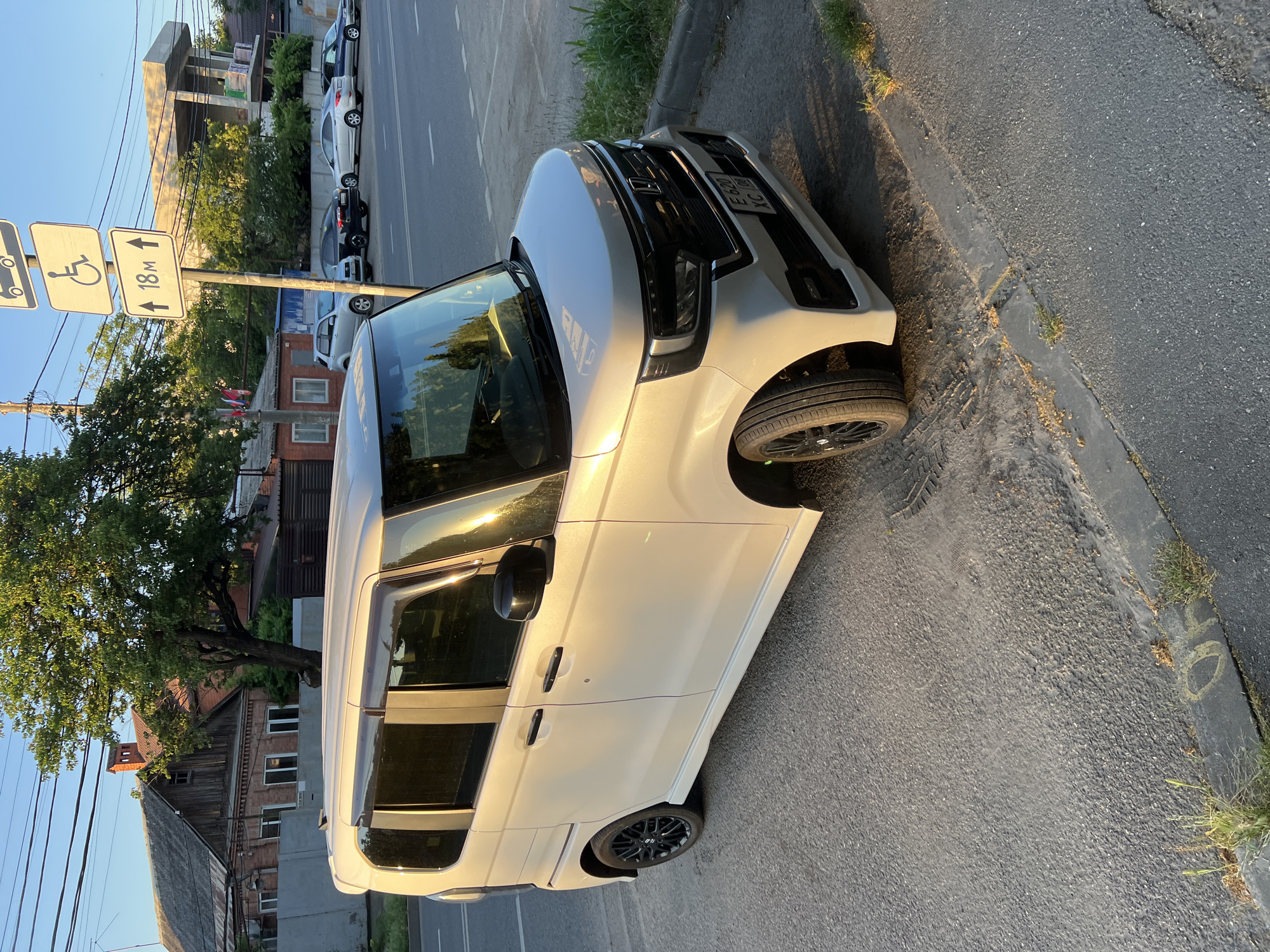 The image depicts a white Honda N-BOX Kei car parked on the side of a street in Krasnodar, Russia. The car is positioned at an angle, facing towards the right of the frame. The street is paved with asphalt and a concrete curb runs along its edge. Beyond the curb, there is a patch of dry grass and dirt. In the background, several other cars are parked in a row. To the left of the street, there are residential buildings and trees. To the right, a modern building under construction is visible, along with traffic signs. The sun is low in the sky, casting long shadows and a warm light on the scene. One of the traffic signs indicates "18m" with arrows pointing in both directions, and another displays a wheelchair symbol, suggesting a parking area for people with disabilities. The license plate of the car is visible and reads "E 620 XC 06".