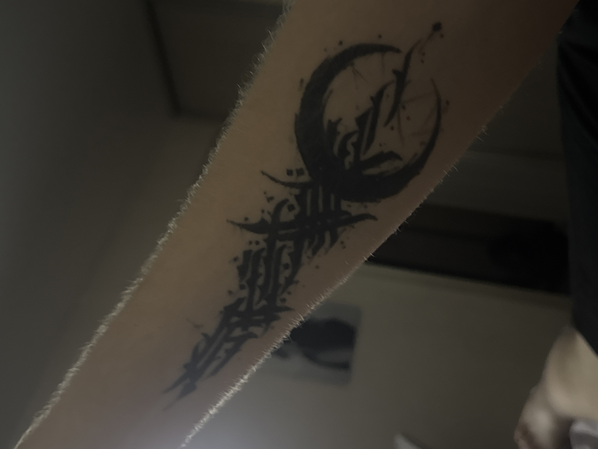 The image shows a close-up of a black tattoo on a person's arm. The tattoo is abstract and appears to be a combination of sharp, angular shapes and a crescent moon or circular element. The arm has light-colored hair. The background is dark and out of focus, with hints of a room containing muted colors and possibly some furniture. There is no discernible information in the image to suggest a location in Saratov, Russia.
