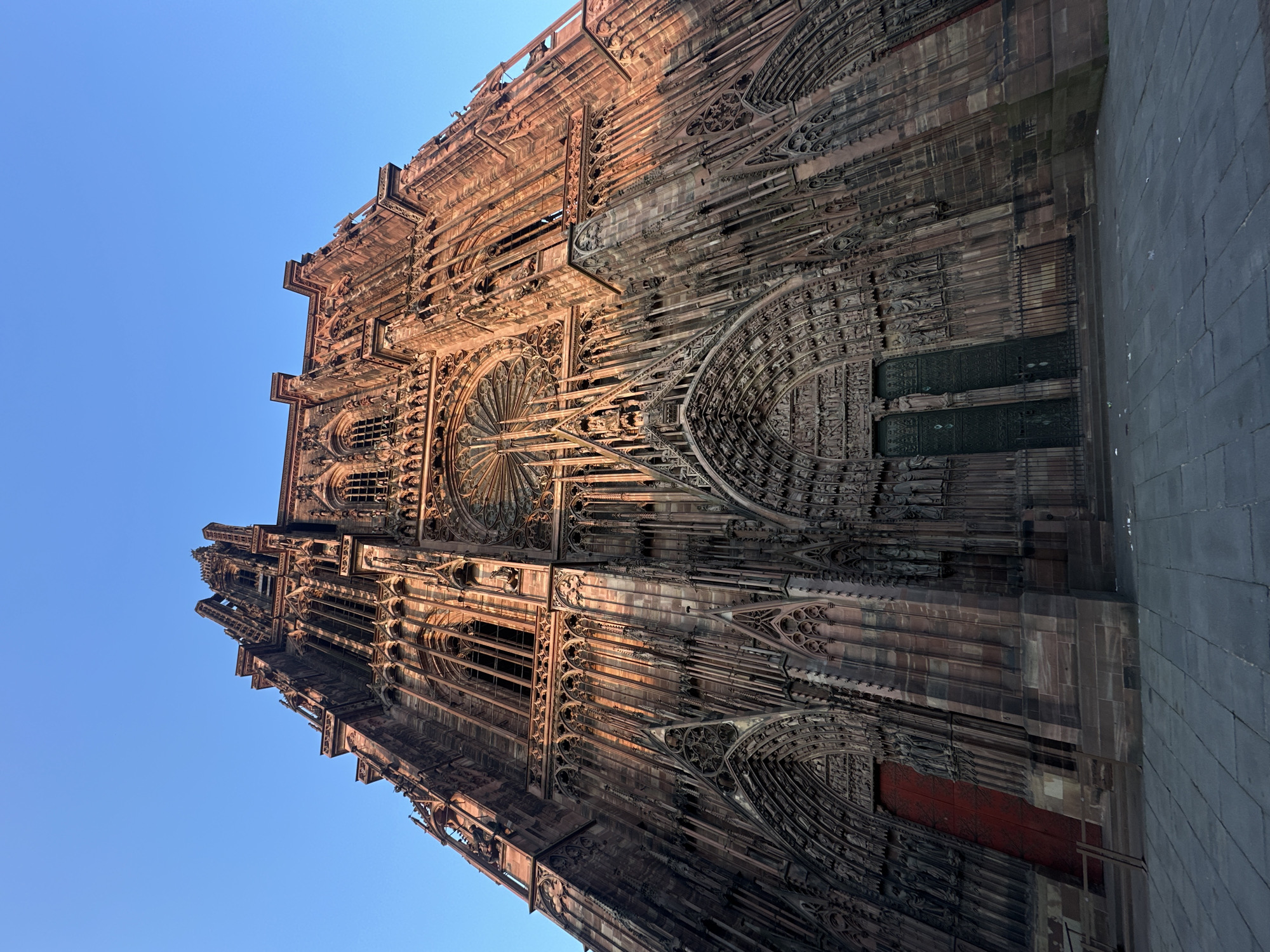 The image shows a low-angle, upward view of the Strasbourg Cathedral (Cathédrale Notre-Dame de Strasbourg) in Strasbourg, France. The cathedral's Gothic architecture is evident in its intricate stone facade, including detailed carvings, arches, windows, and spires. The building is constructed from pink sandstone. The sky behind the cathedral is a clear blue. To the right, a section of the paved ground surrounding the cathedral is visible. The image orientation is tilted.