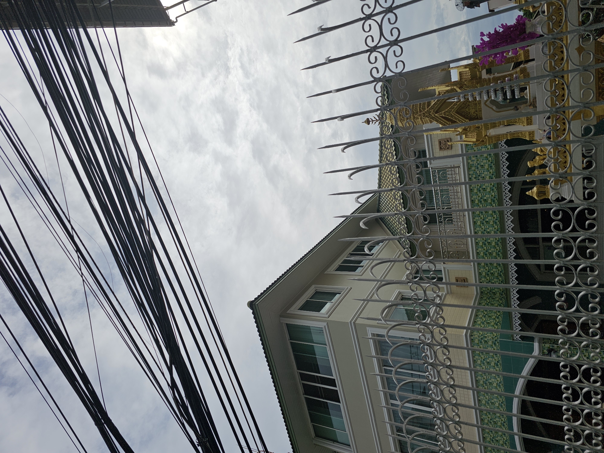 The image depicts a street view in Bangkok, Thailand, looking upwards towards a cloudy sky interlaced with a dense network of black electrical wires. In the foreground, a decorative metal fence with pointed finials and ornate scrollwork partially obstructs the view. Behind the fence, a light-colored, multi-story building with a green tiled roof is visible. The building features numerous windows, some with reflective green glass. The lower section of the building is adorned with green and light green mosaic tiles. To the right, a small, elaborate gold shrine is partially visible, decorated with intricate details and topped with a tiered silver spire. A cluster of purple flowers in a clear vase sits next to the shrine, also behind the fence. The overall scene suggests a residential or commercial area with overhead utility infrastructure common in Bangkok.