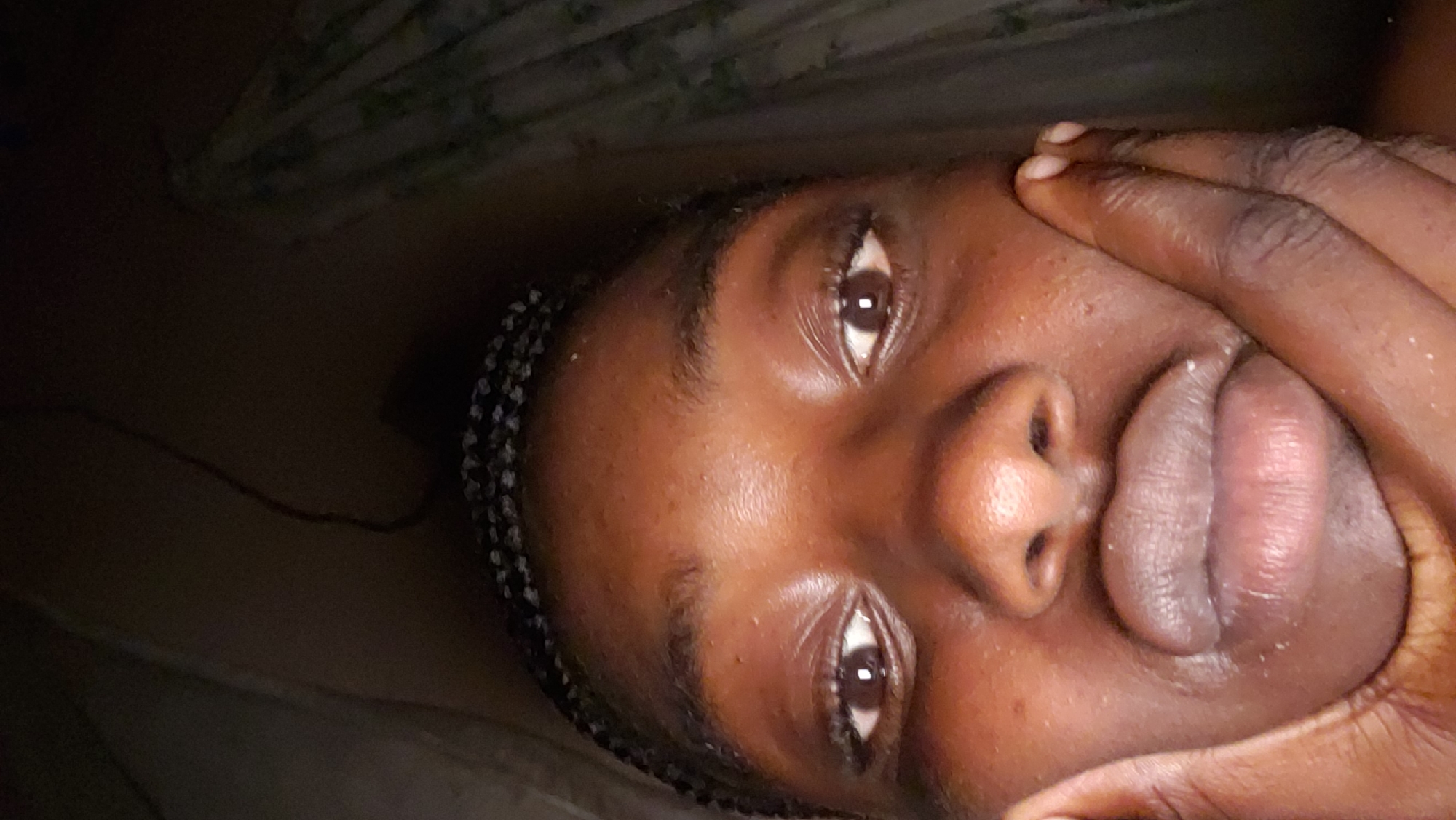 The image presents a close-up, low-angle portrait of an individual with dark skin and dark braided hair along the hairline. The subject's eyes are open, with visible dark irises, and gaze directly at the viewer. The mouth is intentionally pursed, creating a prominent, rounded lip shape. A hand with dark skin is pressed against the subject's left cheek (viewer's left), with fingers extending towards the temple area. The scene is dimly lit, with a dark, indistinct background. A faint vertical pattern, potentially a curtain or draped fabric, is visible on the right side of the background, and a thin, dark line, possibly a wire or cord, appears in the upper central background.
