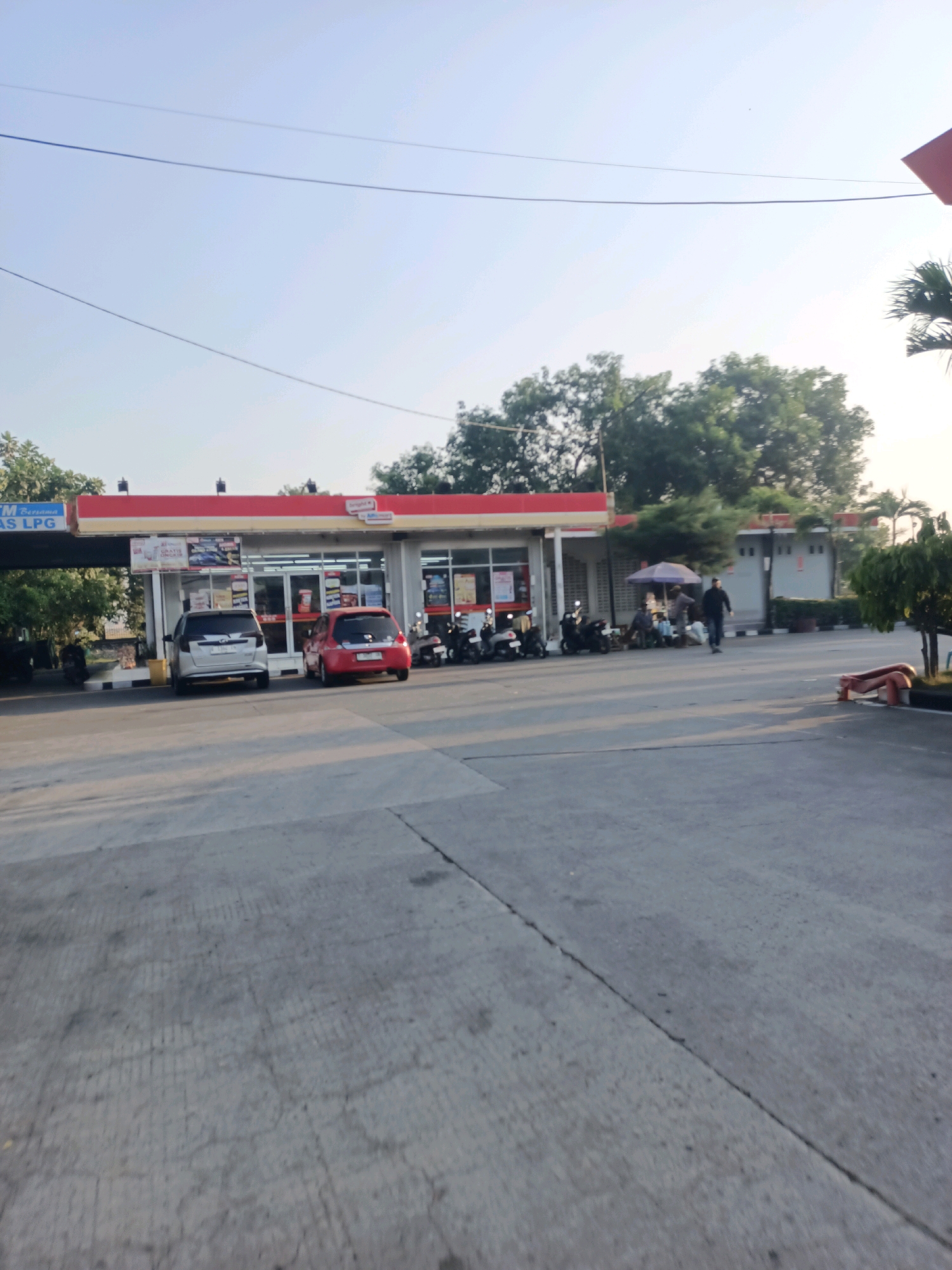 The image depicts an exterior view of a gas station and convenience store in Subang, Indonesia. In the foreground, a concrete paved area shows tire marks. A white SUV and a red hatchback are parked in front of the convenience store. To the left, a sign reads "GAS LPG." Several motorcycles are parked to the side of the store. In the mid-ground, a few individuals are present near a small stall under a purple umbrella, and one person is walking away from the camera. The background features trees and a clear sky with visible overhead wires. The architecture of the convenience store includes a red awning and glass storefronts. The overall impression is a typical daytime scene at a roadside convenience store.