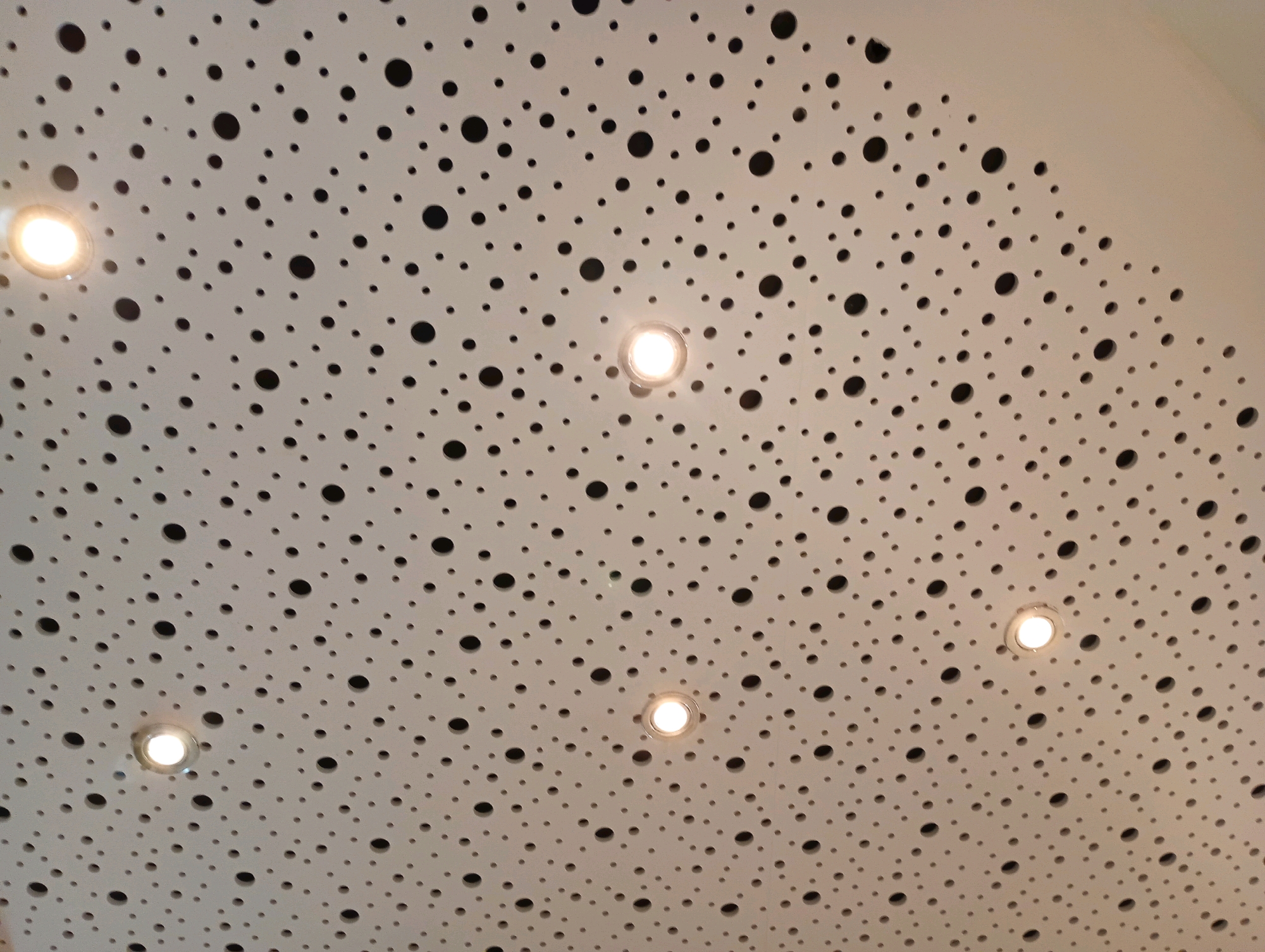 This image shows a close-up, angled view of a white, perforated ceiling. The ceiling has a repeating pattern of numerous circular holes of varying sizes, with some larger holes interspersed among smaller ones. Several recessed circular light fixtures are visible, emitting a warm, white light. The lighting suggests an indoor environment, possibly a modern public space or a commercial building. There are no people or specific identifying landmarks visible in the image that would indicate its location within Bad Sachsa, Germany.
