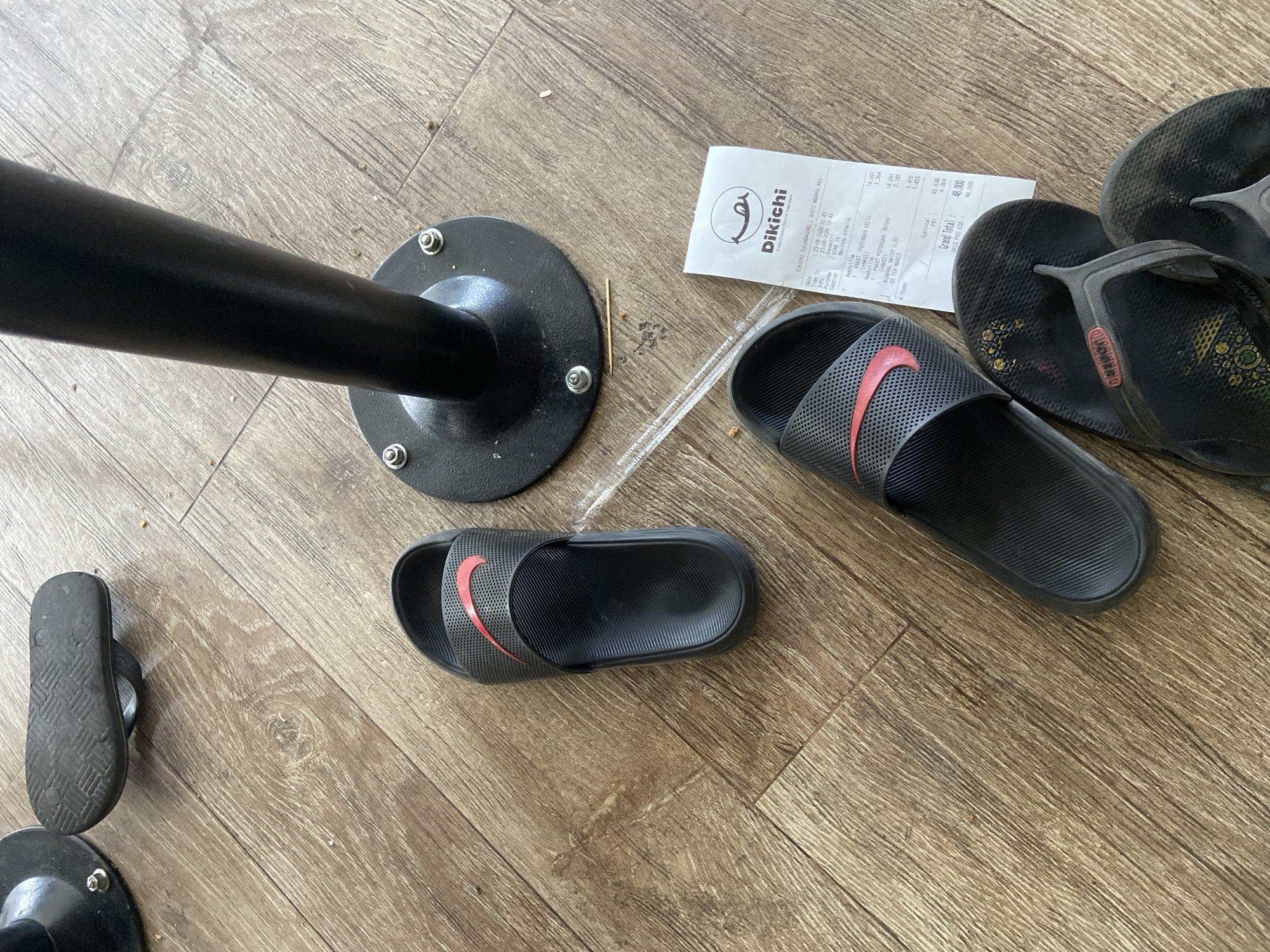 A receipt from Dikichi fried chicken is lying on a wooden floor next to several pairs of black sandals. The receipt, dated 23-08-2020, shows an order from Tulungagung I Gusti Ngurah Rai, Indonesia. The items purchased include "PAKET POTONGAN KECIL (NAKESI)" and "PAKET POTONGAN BESAR (NAKESI)". The total cost is 48,000. A clear plastic straw is also on the floor. The scene appears to be indoors, possibly a dining area.