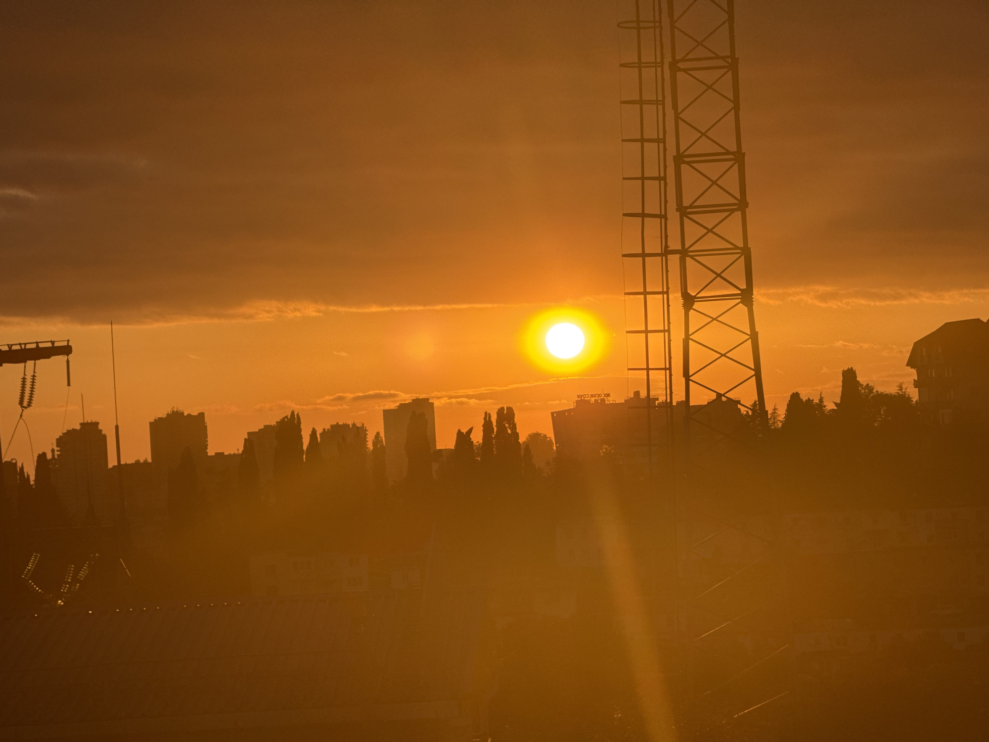 The image shows a sunset over the city of Sochi, Russia. The sun is a bright orange ball in the sky, surrounded by a hazy orange glow. Silhouettes of buildings and trees are visible against the sunset. A tall metal structure, possibly a radio tower, is prominent in the foreground. The scene is bathed in the warm light of the setting sun, creating a dramatic and atmospheric effect.