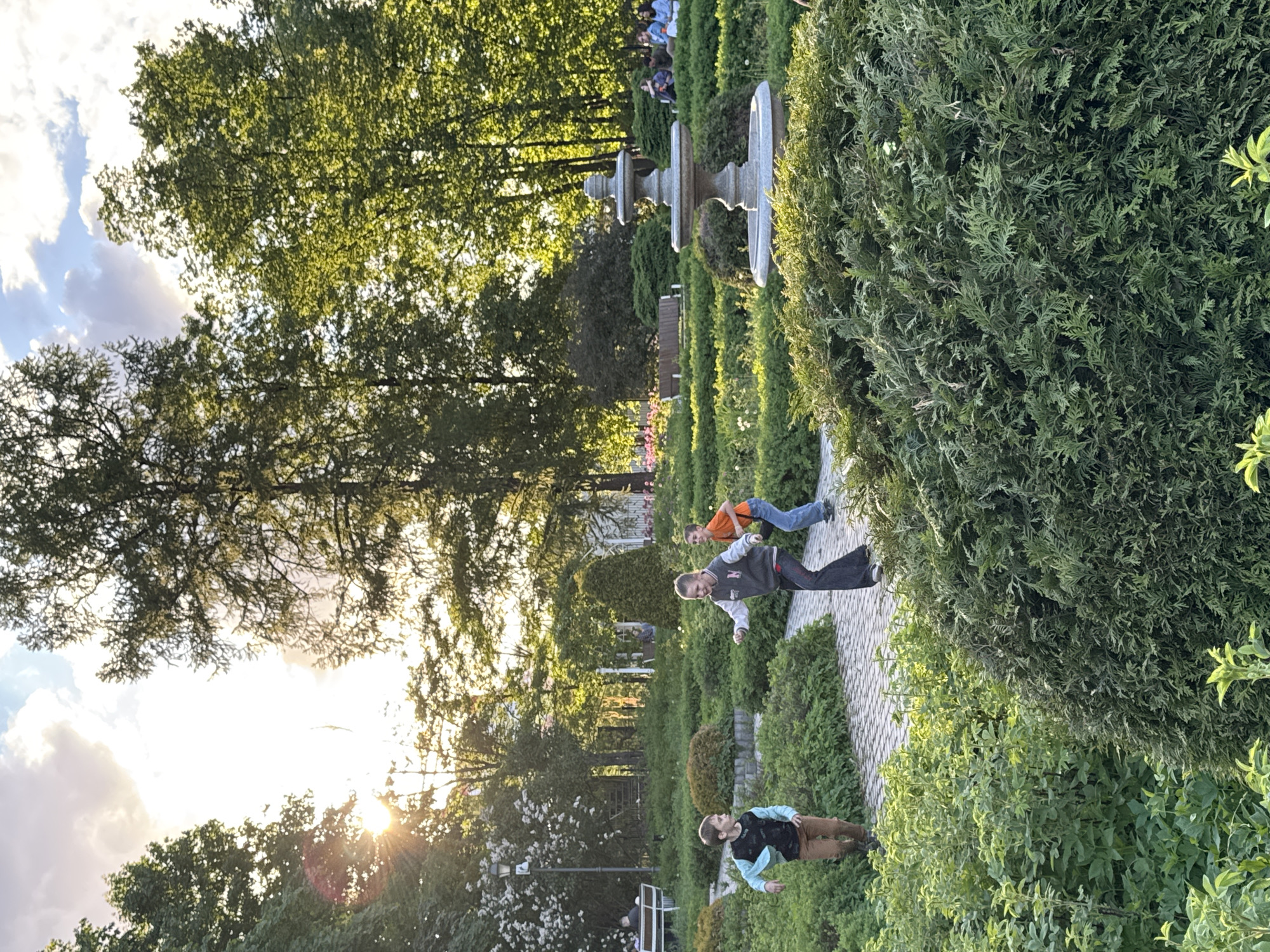 The image depicts an outdoor scene in a garden or park, likely located in Moscow, Russia, during the late afternoon or early evening based on the lighting. Three young boys are seen along a stone-paved walkway winding through manicured bushes and trees. One boy is wearing brown pants and a blue/black shirt and appears to be walking away from the camera. Another boy, wearing dark-colored pants and a gray and white jacket, is leaping or jumping. Further along the path, the third boy, clad in an orange shirt and jeans, appears to be running or leaping.
The environment includes lush green vegetation, trimmed bushes, and tall trees. A decorative fountain stands in the background, with people seated on benches in the distance beyond the fountain. The sky is visible through the trees, showing the sun and some clouds. The scene is generally tranquil and bathed in golden sunlight.