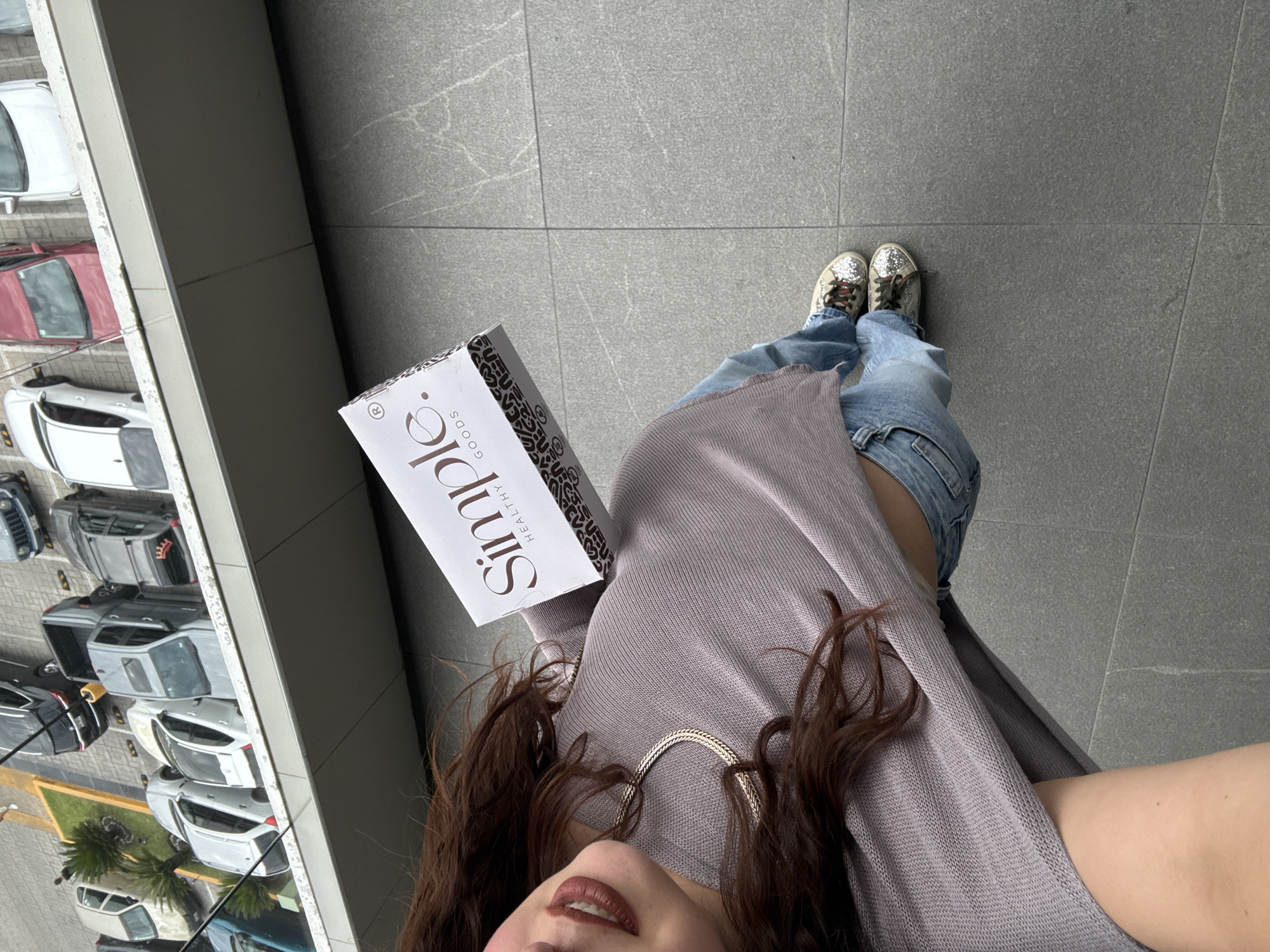 In San Bernardino Tlaxcalancingo, Mexico, a person is viewed from a high angle, holding a white box from "Simple. HEALTHY GOODS" in their right hand. The individual wears a light purple, loose-fitting top, light blue jeans, and sparkly sneakers. Their brown hair is visible, and their face, with dark lipstick, is turned to the left. The background consists of a gray tiled surface and an elevated parking lot containing several cars, including a black Jeep, a red sedan, and various other vehicles.