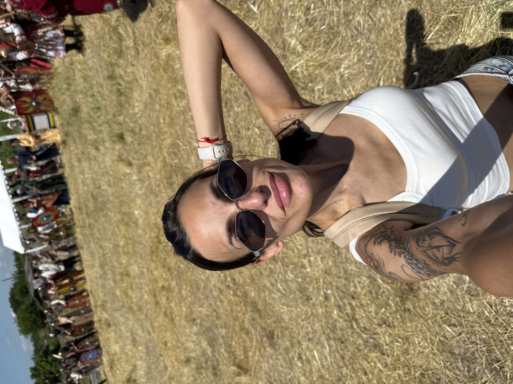 The image captures a female subject taking a selfie in a sunlit, open field in Gigen, Bulgaria. The woman in the foreground has dark hair pulled back, wears hexagonal-framed sunglasses, a white tank top, and light-colored shoulder straps. Visible tattoos include an intricate design on her upper left arm and a stylized sun on her upper right arm. A light-colored watch and a red string bracelet are present on her right wrist. She displays a slight smile. The immediate ground consists of dry, golden-brown grass. In the blurred background, a historical reenactment is underway, featuring a line of individuals. Many are dressed in Roman-era military attire, including tunics, armor, and shields with various painted designs. Other participants wear historical civilian clothing. A white tent structure is visible on the right side of the background, with distant trees under a blue sky with sparse clouds.