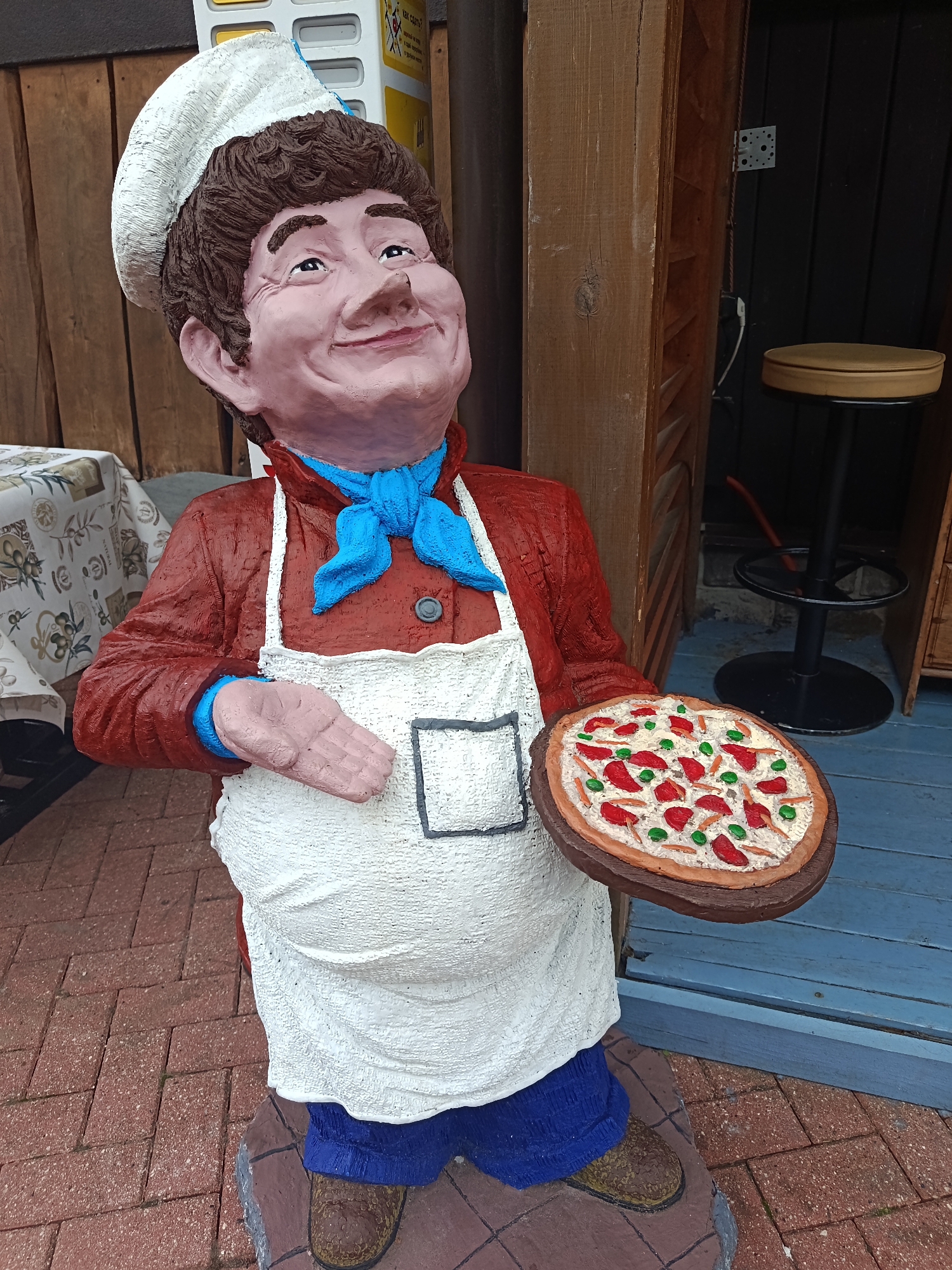 A full-shot, eye-level, outdoor, natural light photograph captures a weathered, life-size statue of a smiling chef in Estosadok, Russia, holding a pizza. The chef, with rosy cheeks, wide eyes, and a brown, textured wig, wears a white chef's hat, a red jacket with a blue ascot, and a white apron with a black-trimmed pocket. His right hand, with visible fingers, is extended forward, while his left hand holds a small, circular pizza with red toppings and green garnishes. The statue stands on a red brick pathway, adjacent to a wooden-clad building with blue-painted steps. In the background, a rustic table with a patterned tablecloth is visible to the left, and a brown bar stool with a metal base is positioned to the right, partially obscured by a wooden beam. The overall scene suggests a welcoming atmosphere at a food establishment.