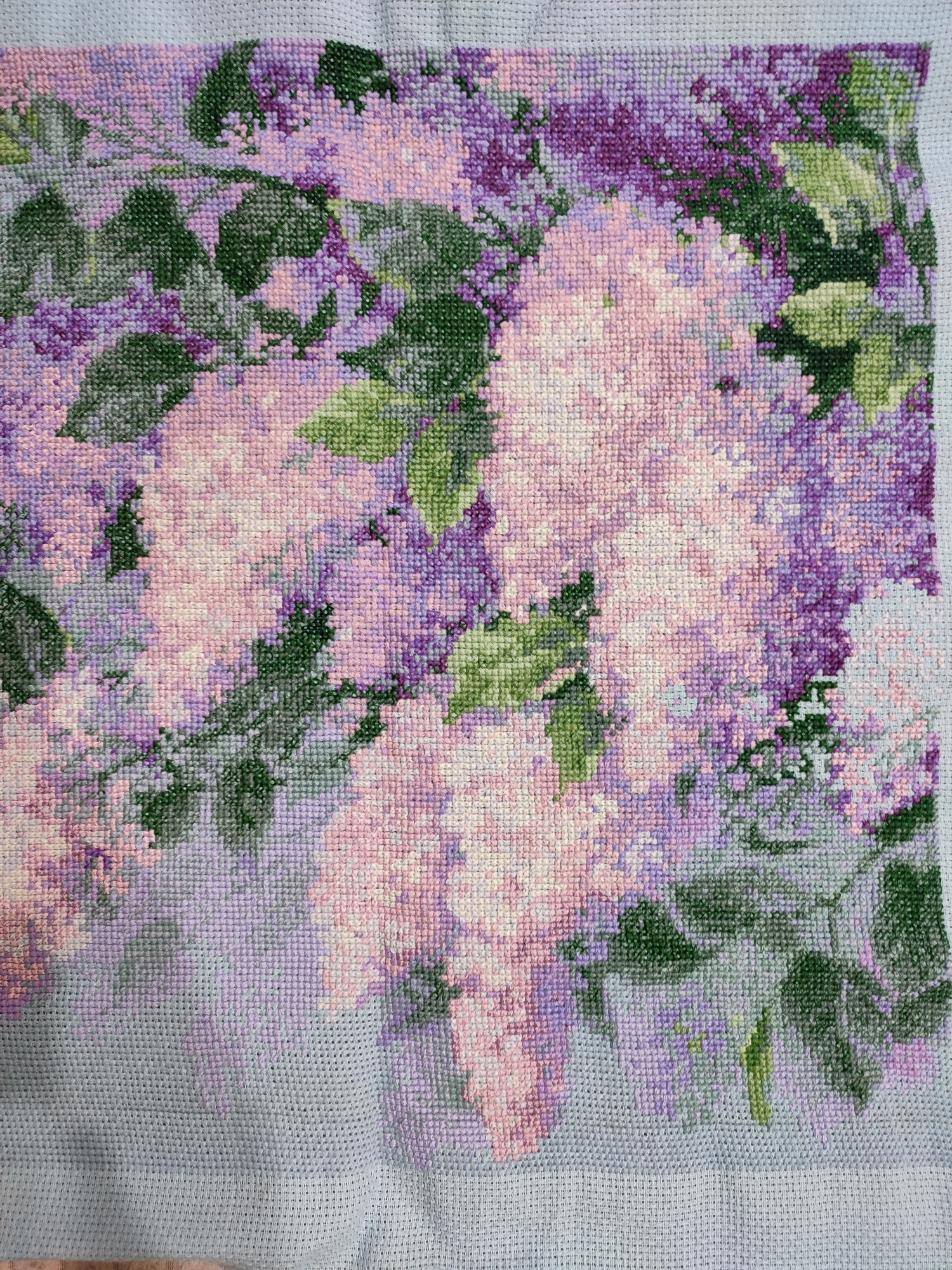 This is a close-up, overhead shot of a cross-stitch project depicting a cluster of lilacs in bloom. The lilacs are rendered in various shades of purple, pink, and white, with dark green leaves interspersed throughout. The stitches are small and precise, creating a textured, pixelated effect. The background is a light blue fabric, with the edges of the cross-stitch work visible. The lighting is even and bright, suggesting an indoor setting, possibly a craft room or a well-lit area. There are no people or animals visible in the image. The focus is entirely on the detailed craftsmanship of the cross-stitch art. The image appears to be a photograph of a work in progress, as some of the blue fabric is still uncovered. The composition is tightly cropped, emphasizing the delicate beauty of the flowers and the meticulous nature of the embroidery.