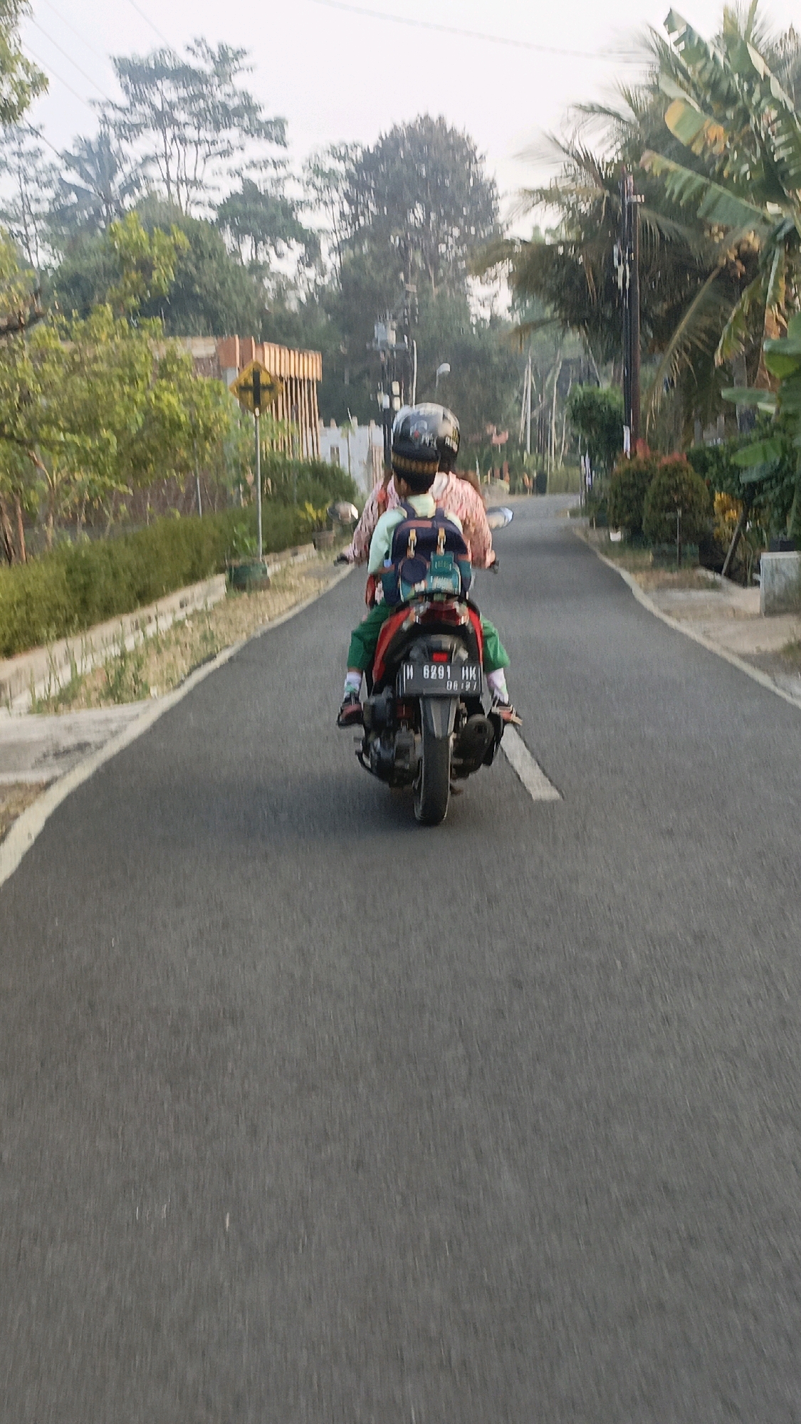 The image shows two individuals riding a motorcycle from behind on a paved road in Salatiga, Indonesia. The person in front, who appears to be an adult, is wearing a pink and white patterned shirt and a black helmet. The person in the back, likely a child, is wearing a light green shirt, green pants, and a black helmet. A blue and red backpack is strapped to the back of the child. The motorcycle has a license plate that reads "H 6291 HK 08.27". The road is flanked by a green embankment on the left and vegetation and trees on the right. In the background, there are more trees, some buildings, and utility poles. The sky is overcast. The overall scene depicts a common mode of transportation in a semi-rural or suburban setting in Indonesia.