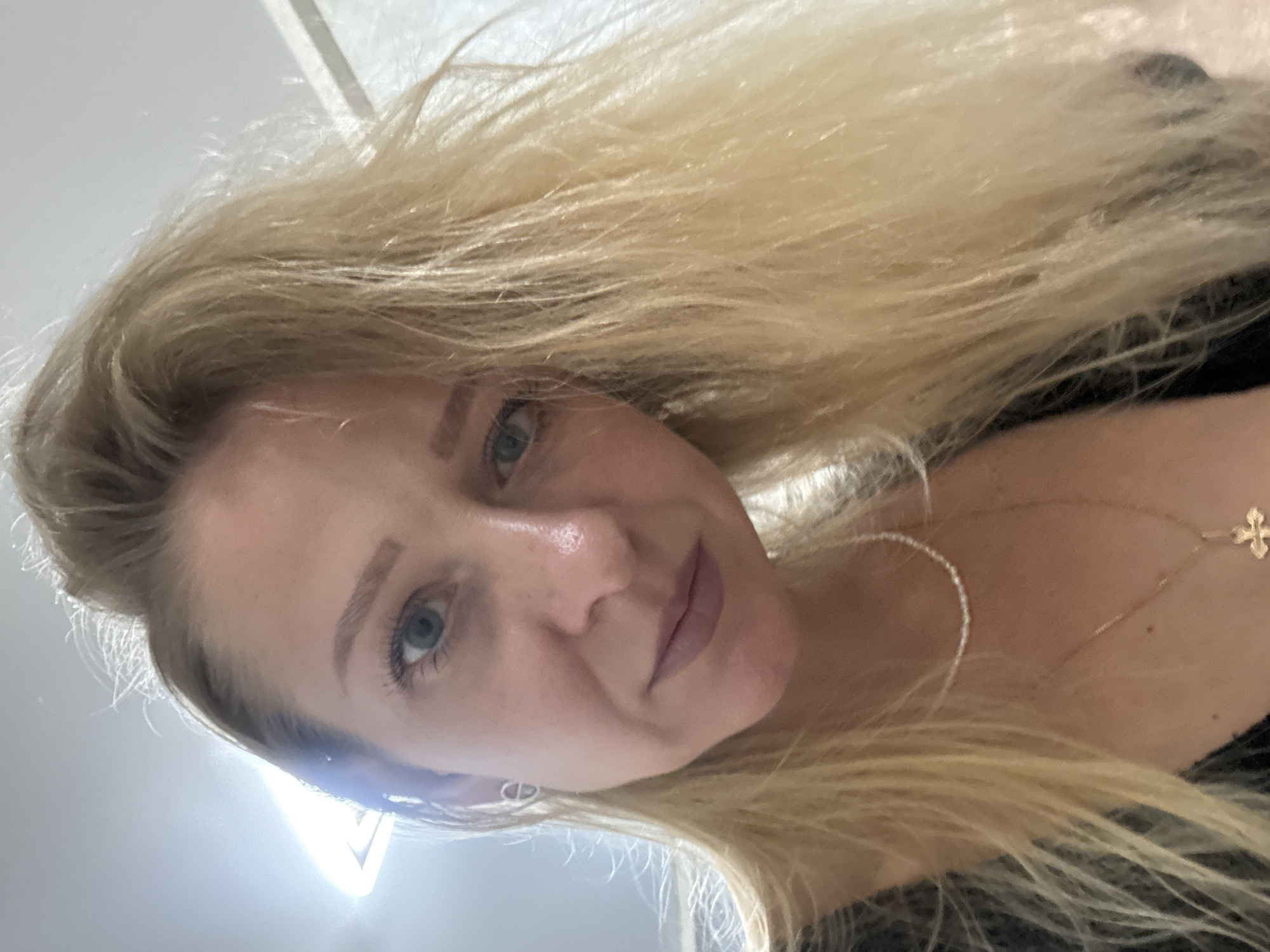A close-up selfie shows a woman with blonde hair and blue eyes, tilted her head and looking directly at the camera. Her hair is a light blonde and appears somewhat tousled. She is wearing makeup, with eyeliner and mascara visible on her eyes, and a matte lipstick on her lips. A delicate necklace with a small cross charm is visible around her neck. The lighting suggests it might be daytime, possibly indoors due to the bright, diffused light source visible in the lower left corner of the image, which creates a lens flare. The background is largely out of focus, but appears to be a neutral-colored wall or ceiling. No text is visible in the image.