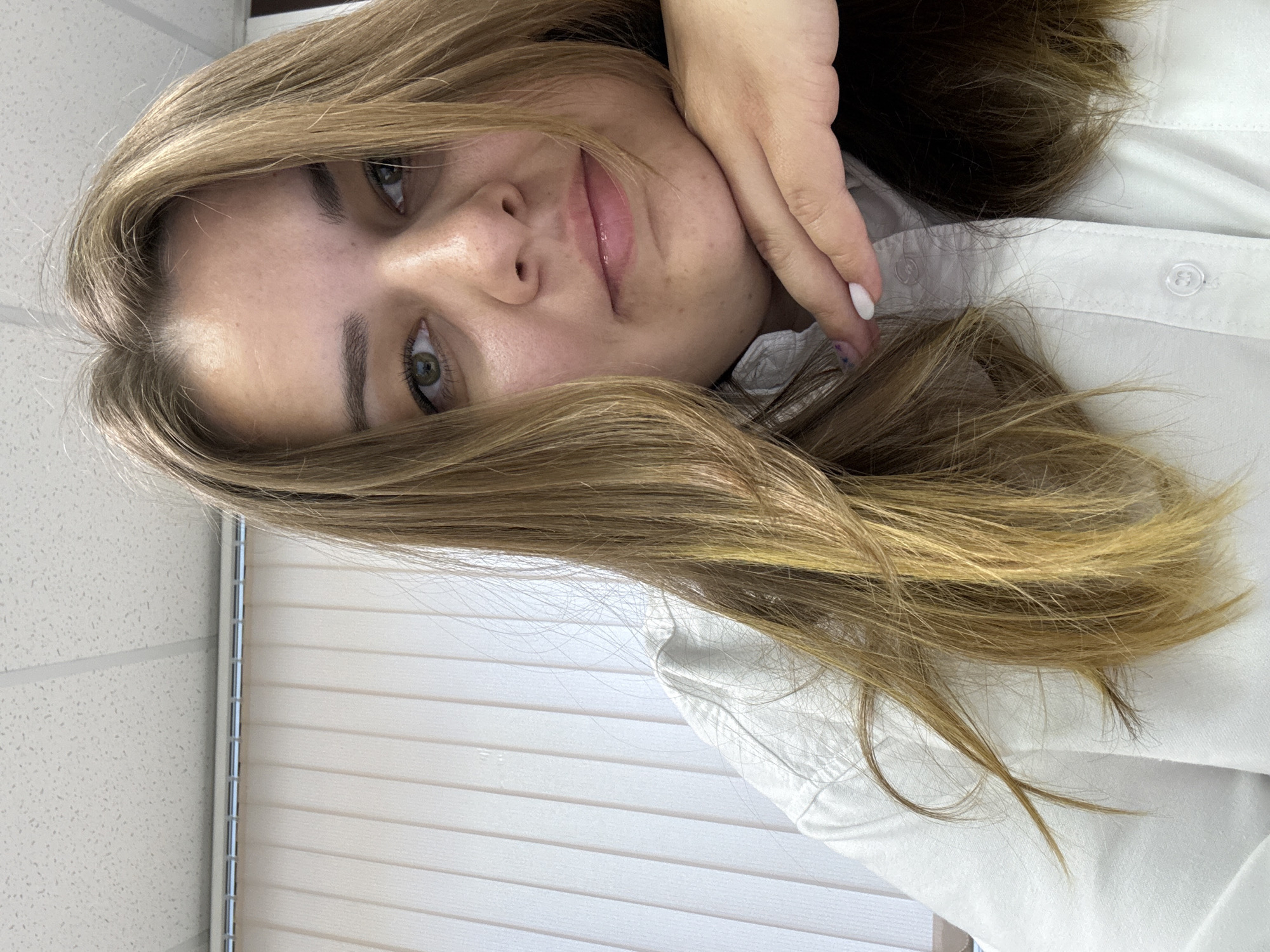A fair-skinned person with light brown hair is shown in a close-up, angled view. The person's face is prominently displayed, with one hand touching their cheek. They have green eyes. The individual is wearing a white collared shirt. In the background, a closed set of white horizontal blinds is visible. There is no definitive evidence of location or specific city.