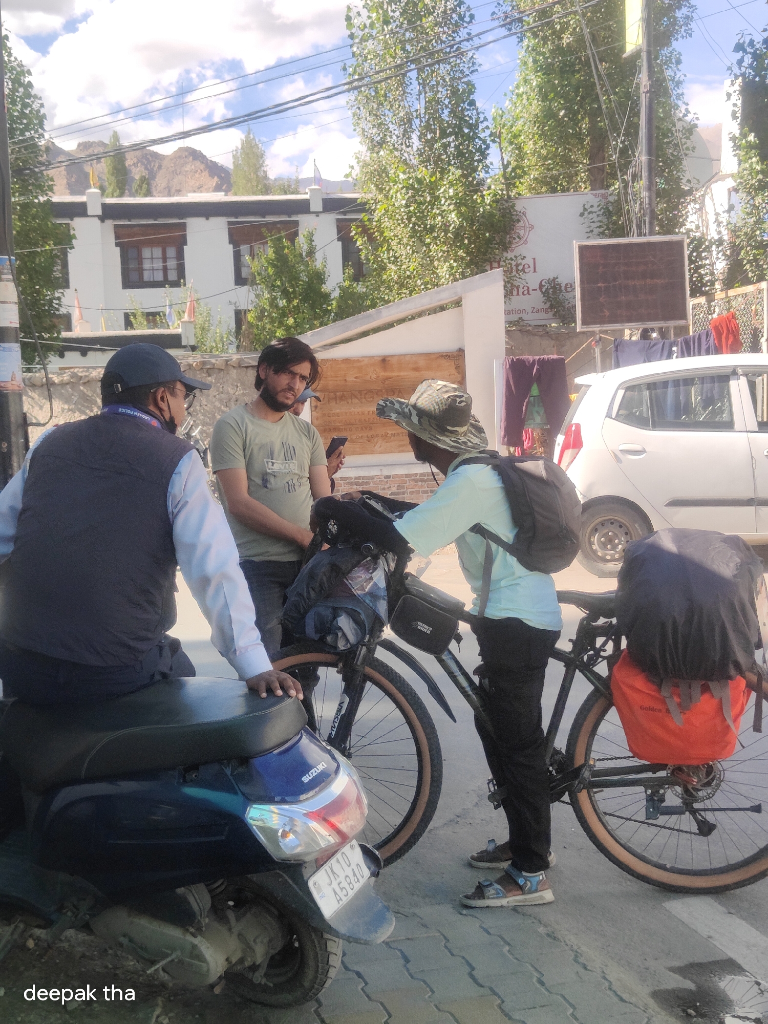 In Leh, India, on a sunlit street, a police officer wearing a dark vest over a light blue shirt and a navy blue cap speaks with two men near a parked scooter and a bicycle laden with luggage. The officer has glasses and a badge clipped to his vest. One man, wearing a light green t-shirt, is looking down at his phone. The other man, wearing a light blue shirt and a camouflage hat, is standing next to his bicycle, which has backpacks attached to the frame and handlebars. A white car is parked to the right of the scene. The background features a building with white walls and dark windows, along with trees and mountains. The scooter has a license plate that reads "JK10 A5840". The overall atmosphere suggests a casual interaction or an exchange of information on a street in Leh.