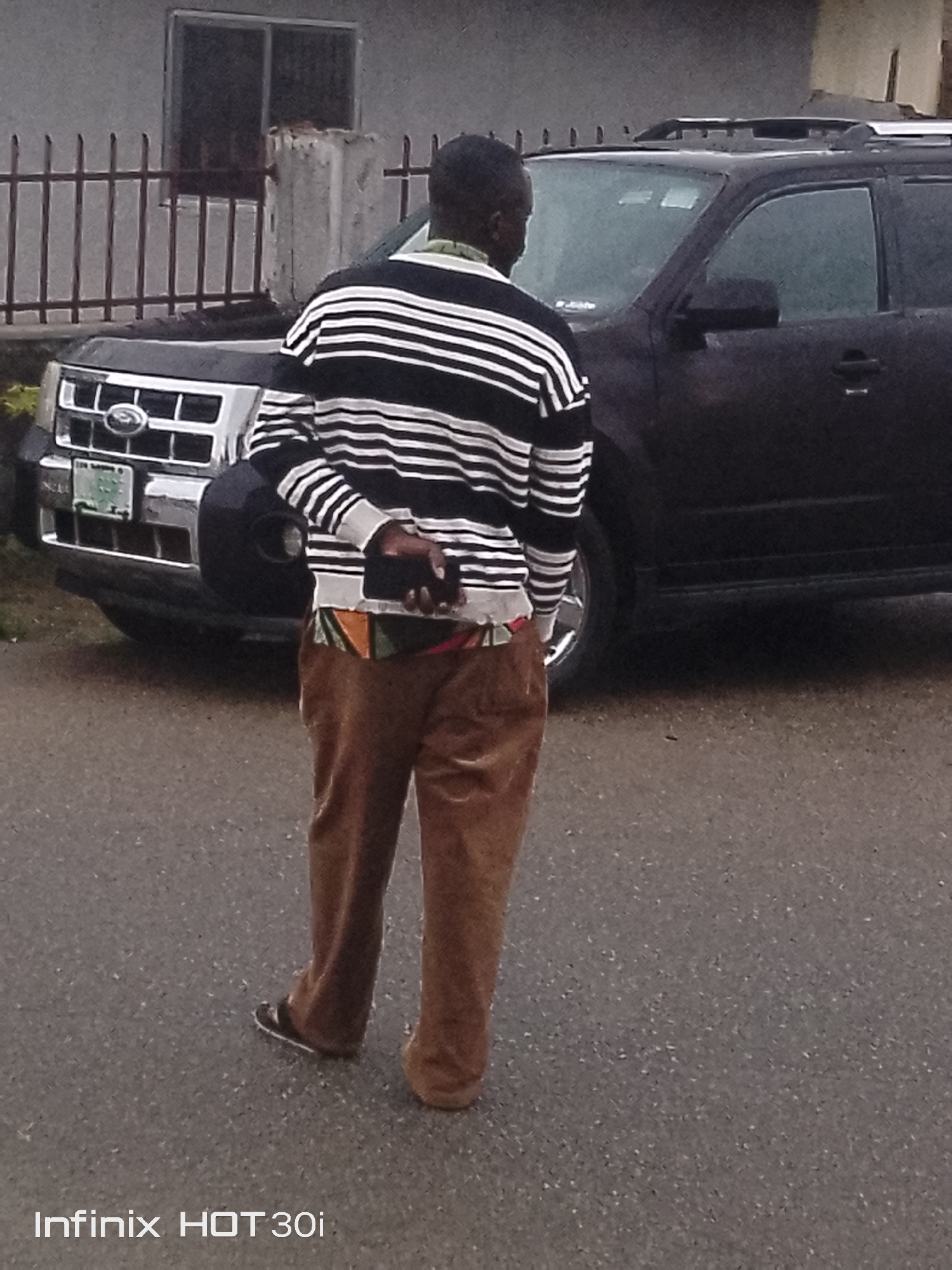 A man stands facing away from the camera, with his left hand behind his back holding a dark object, likely a phone. He is wearing a black and white horizontally striped long-sleeved shirt over a collared shirt, and brown pants. A colorful patterned waistband is visible. He is wearing black sandals. Behind him is a black SUV. A metal fence runs along the side of a building. The ground is paved. The image is taken outdoors in what appears to be an urban or suburban setting.