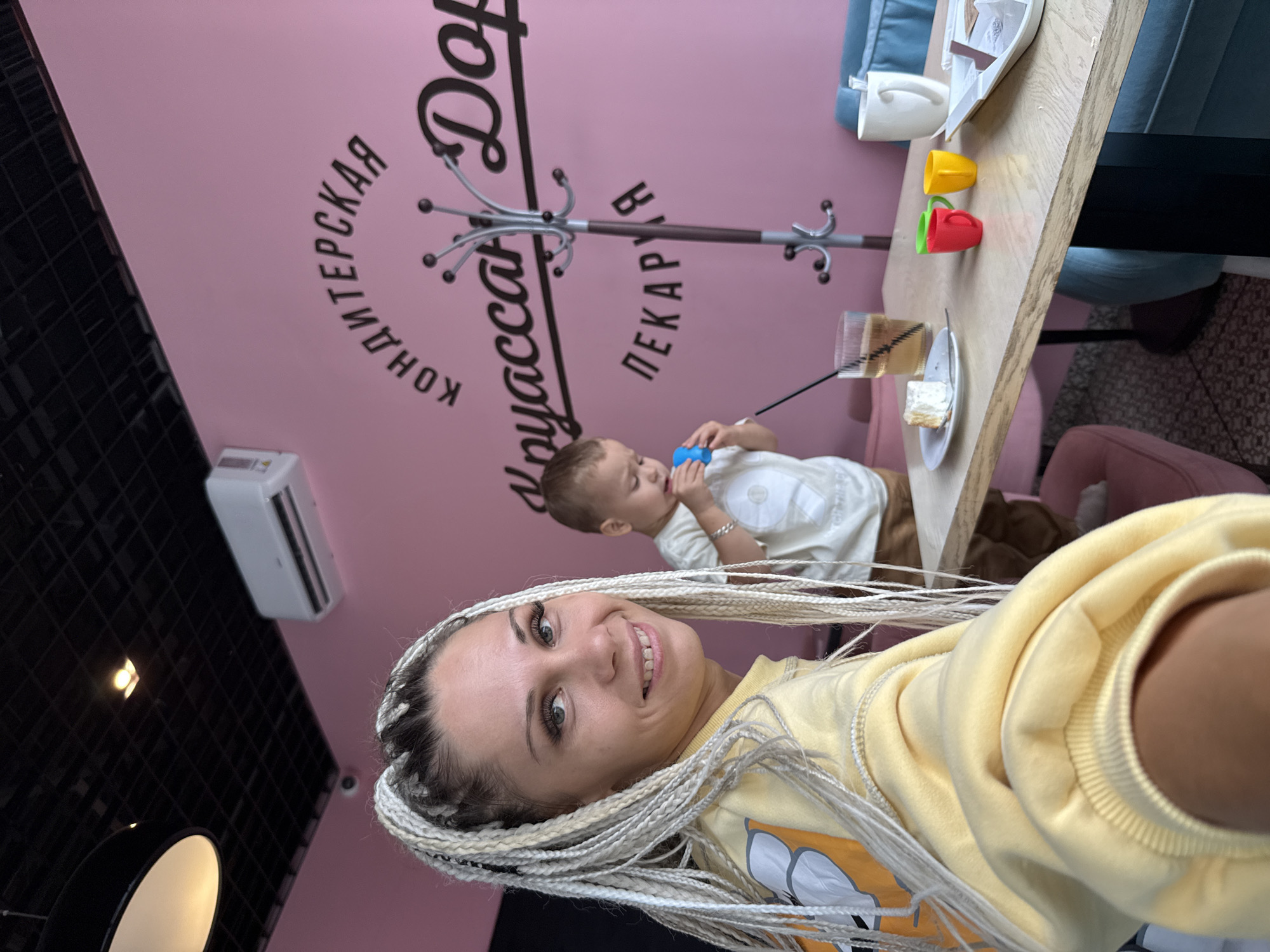 The image depicts an indoor scene, likely a cafe or bakery, with a woman and a child. The woman, with blonde braided hair, is in the foreground, looking at the camera and smiling. She is wearing a yellow sweatshirt. A young child, wearing a white t-shirt, is in the midground playing with a blue toy. A table in the foreground has a slice of cake, a glass of drink, a fork, and three small colorful cups. A coat rack stands behind the table. The walls are painted pink and feature text in black lettering, which appears to be in Russian and translates to "Confectionery Dobro-Pekarnya" (Confectionery Dobro-Bakery). The location is identified as Rusanovo, Russia. The environment appears to be a casual dining or seating area.