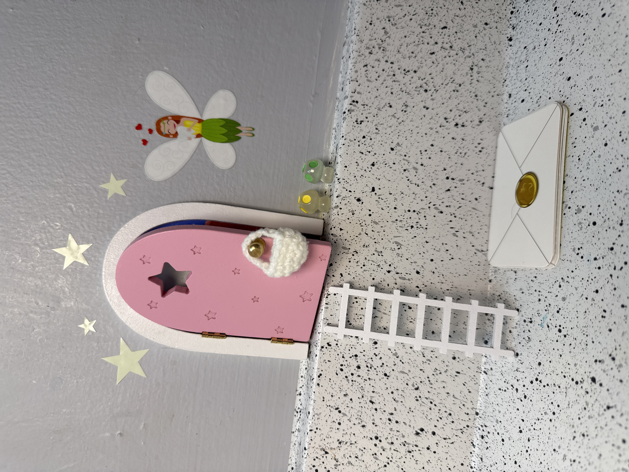 A whimsical fairy door is installed in a room in Columbus, Ohio, United States of America. The door is pink, with a white frame and gold hinges, featuring star cutouts, and is adorned with a white crocheted door knocker that includes a gold bead. A small white ladder leans against the speckled wall leading up to the door. On the wall to the right of the ladder is a white envelope with a gold seal. To the left of the door, there is a sticker of a winged fairy and a scattering of star stickers. Two small glass marbles sit on top of the door frame. The background wall colors are light gray on the left and speckled white on the right.