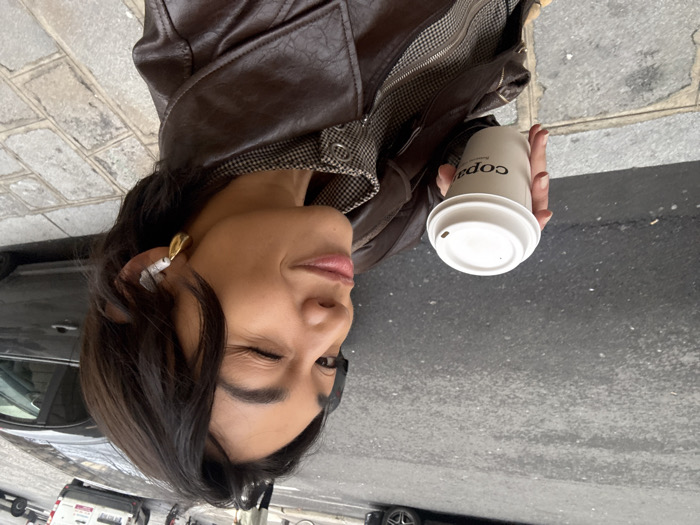 The image depicts a close-up of an adult female standing on a city sidewalk. She has short, dark hair, looks directly at the camera, winks her right eye, and exhibits a subtle smile. She is wearing a dark brown leather jacket over a garment with a small checked or plaid pattern. A white earbud is inserted into her right ear, accompanied by a gold-toned, curved earring. Her left hand firmly grasps a white disposable coffee cup with a white plastic lid, which displays "copa" and "Paris" branding. The immediate foreground features a cobblestone sidewalk. Beyond the sidewalk, an asphalt street is visible, with parked vehicles, including a dark-colored sedan and a white van, and a blurred pedestrian in the background. The location is identified as Paris, France, based on the branding present on the coffee cup.