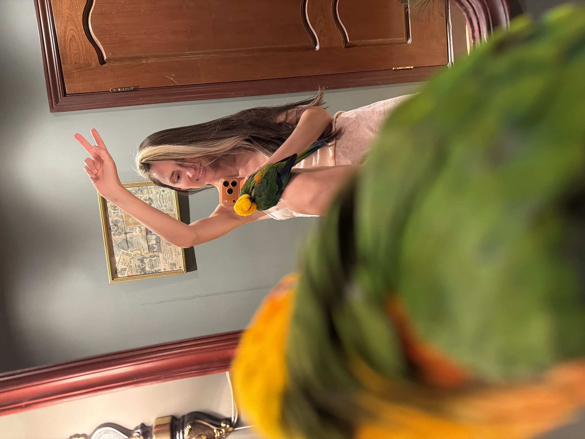 A woman is taking a selfie with her phone while holding a green and yellow parrot. She is making a peace sign with her left hand. A framed picture hangs on the wall behind her. The environment includes a wooden cabinet, a red-trimmed ledge, and a decorative lamp. No location information related to Khimki, Russia is discernable.