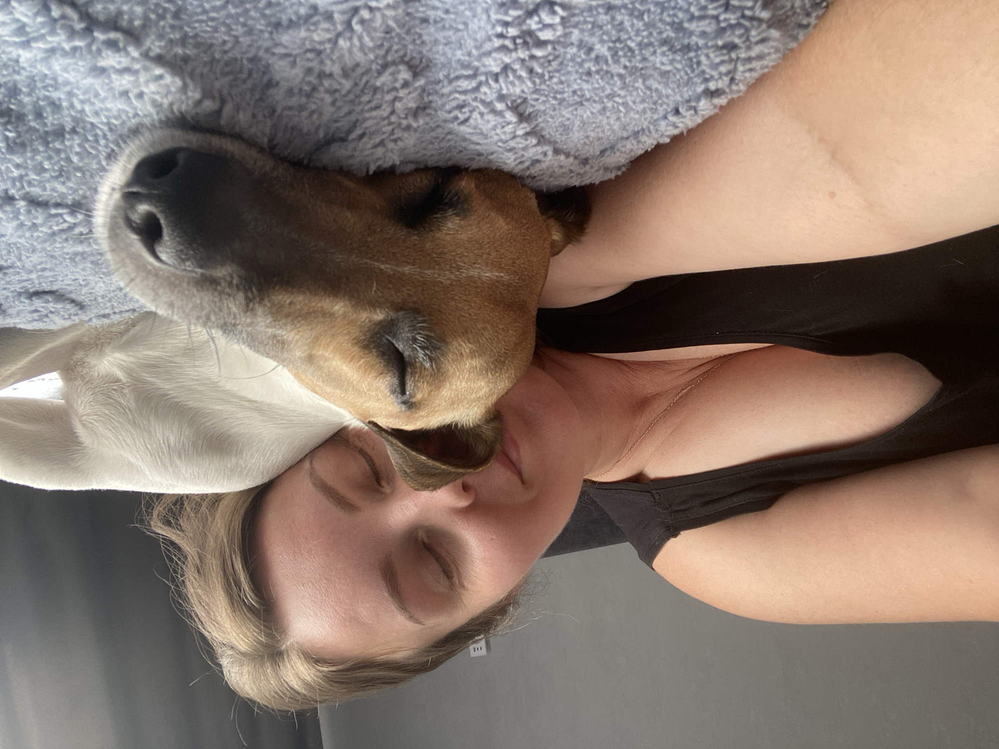 A woman and a dog are sleeping. The woman is on her side, with her head resting on her arm. Her eyes are closed and she has a slight smile on her face. The dog is lying next to her, with its head resting on her shoulder. The dog's eyes are also closed. They are both on a blue, fluffy blanket. The background is a gray wall. The image appears to have been taken indoors, likely in a home. There is no information about the location in the city of Noviy, Russia.