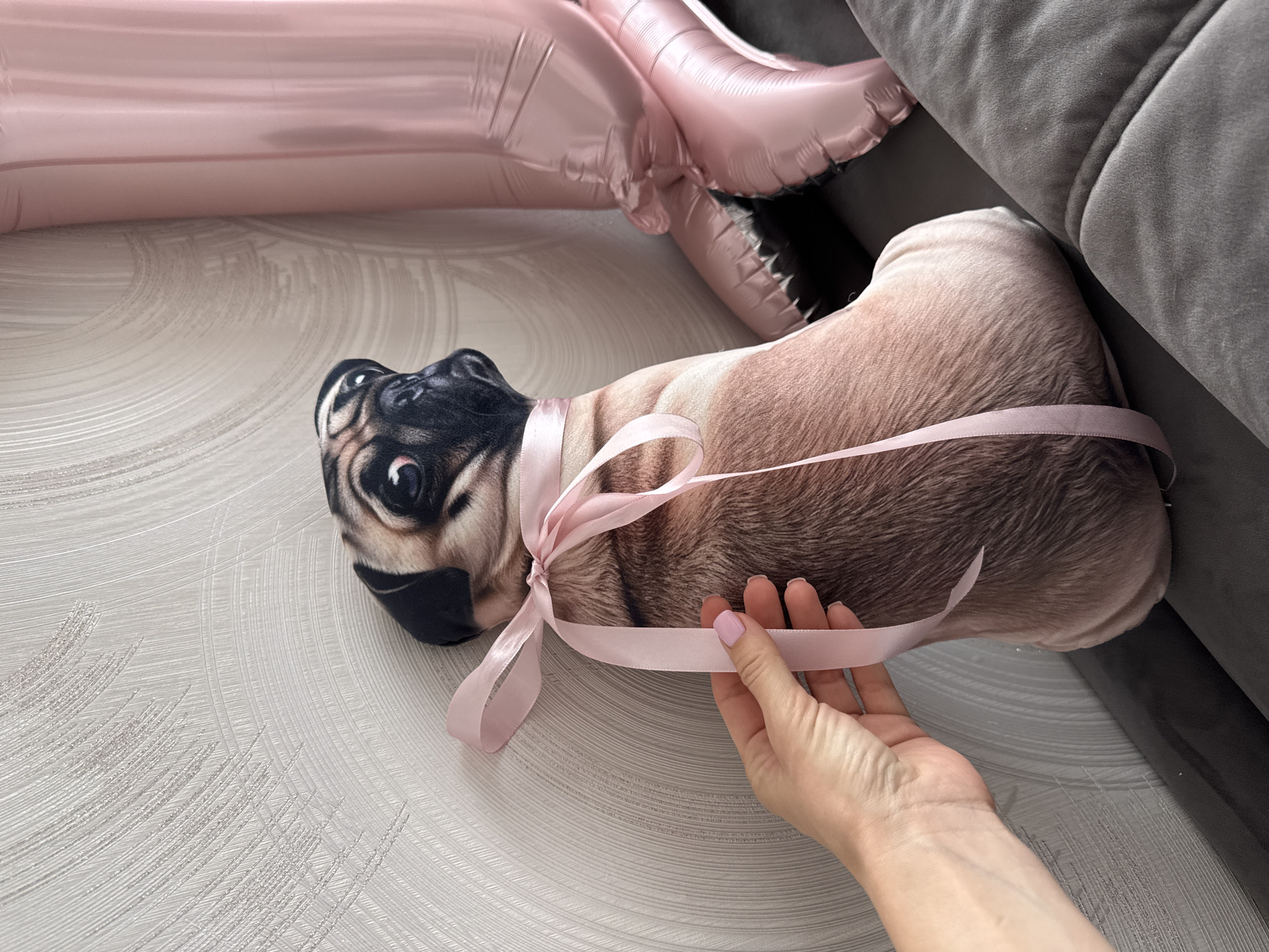 The image presents a still life featuring a pug-shaped pillow tied with a pink satin ribbon. A hand with manicured fingernails is holding the ribbon, drawing attention to the pillow. The backdrop includes a light-colored textured surface, possibly wallpaper or fabric. A pink balloon and a grey sofa provide additional context to the scene. The location, Sochi, Russia, is not visibly discernible from the image's content.