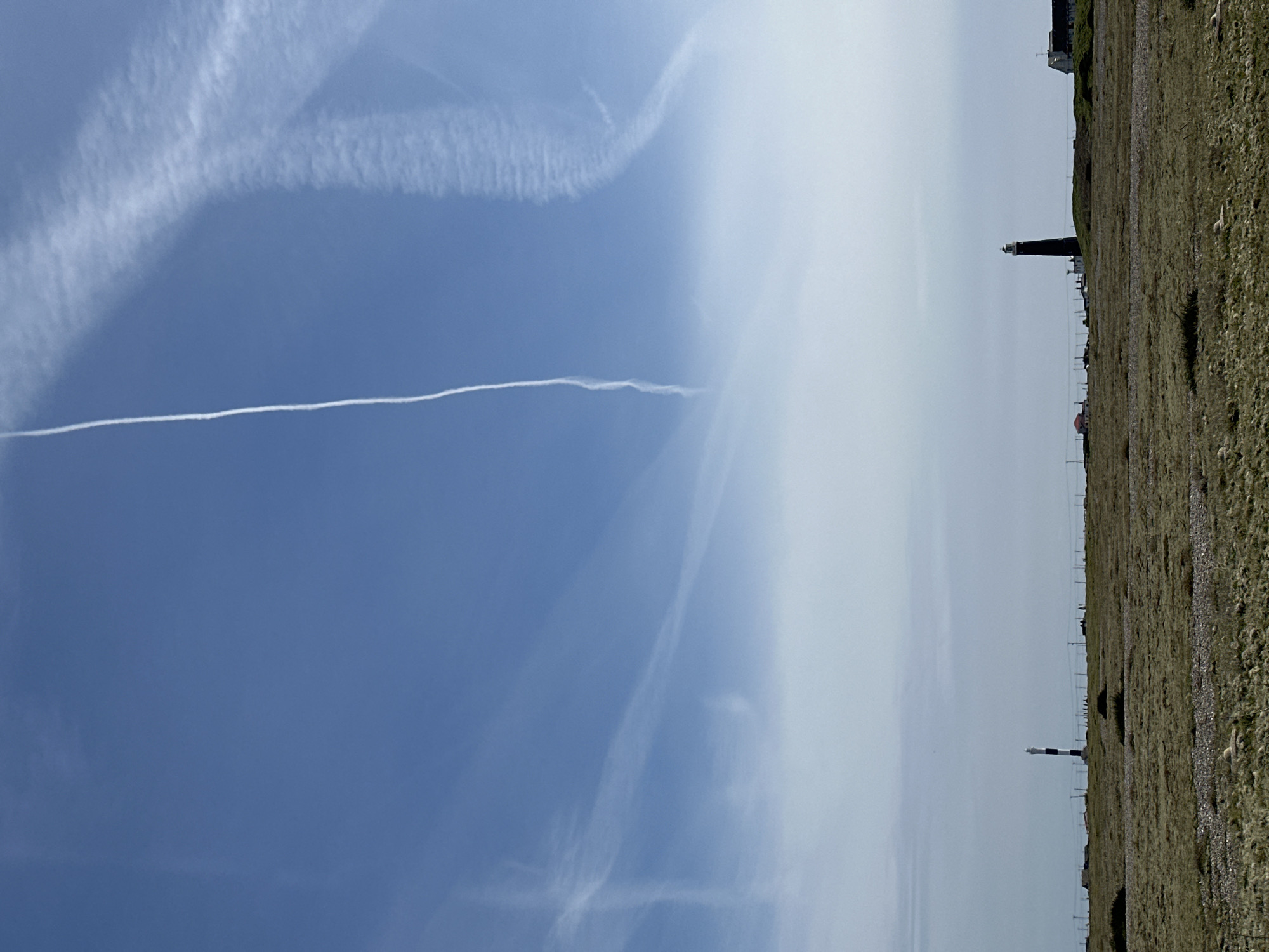 The image depicts a coastal landscape under a blue sky with visible contrails. A thin, straight contrail stretches horizontally across the sky. Other, less defined contrails are present. The land slopes towards the right side of the frame and appears grassy. A lighthouse is present in the distance, along with other structures that appear to be buildings. Power lines stretch horizontally. The location is the city of Folkestone and Hythe District, United Kingdom.