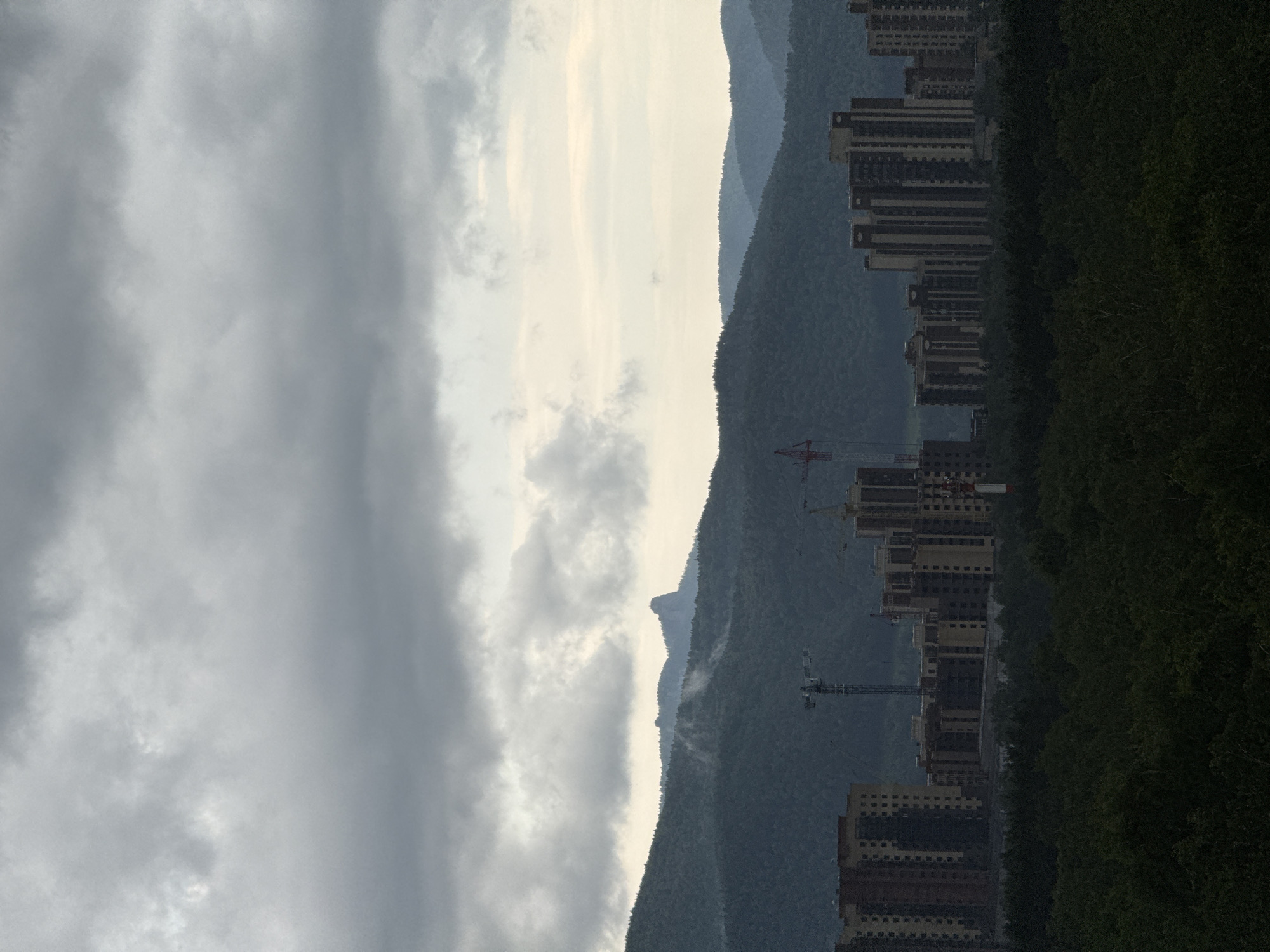 The image depicts a residential area under construction, nestled against a backdrop of densely forested mountains. The perspective is from a higher vantage point, looking down and across the scene. The sky is overcast, with heavy, dark gray clouds dominating the left side of the frame, suggesting a potential for rain or an impending storm. The right side of the frame is filled with layers of dark green trees that form a thick forest.
In the middle ground, multiple tall, multi-story buildings, likely apartment complexes, are visible. Some appear to be completed, while others show signs of ongoing construction, with scaffolding and cranes present. A prominent red and white construction crane is visible, along with other industrial machinery. The buildings are arranged in rows, extending along the base of the mountains. The architectural style suggests modern, high-density housing.
The mountains are a dominant feature, with their slopes covered in vegetation. The hazy atmosphere and cloudy sky create a somewhat somber mood. The time of day is difficult to pinpoint precisely due to the overcast conditions, but the overall lighting suggests it could be late afternoon or early evening. The location context of Krasnoyarsk, Russia, aligns with mountainous terrain and extensive forestation. No visible text is present in the image. The scene conveys a sense of urban development encroaching upon a natural landscape.