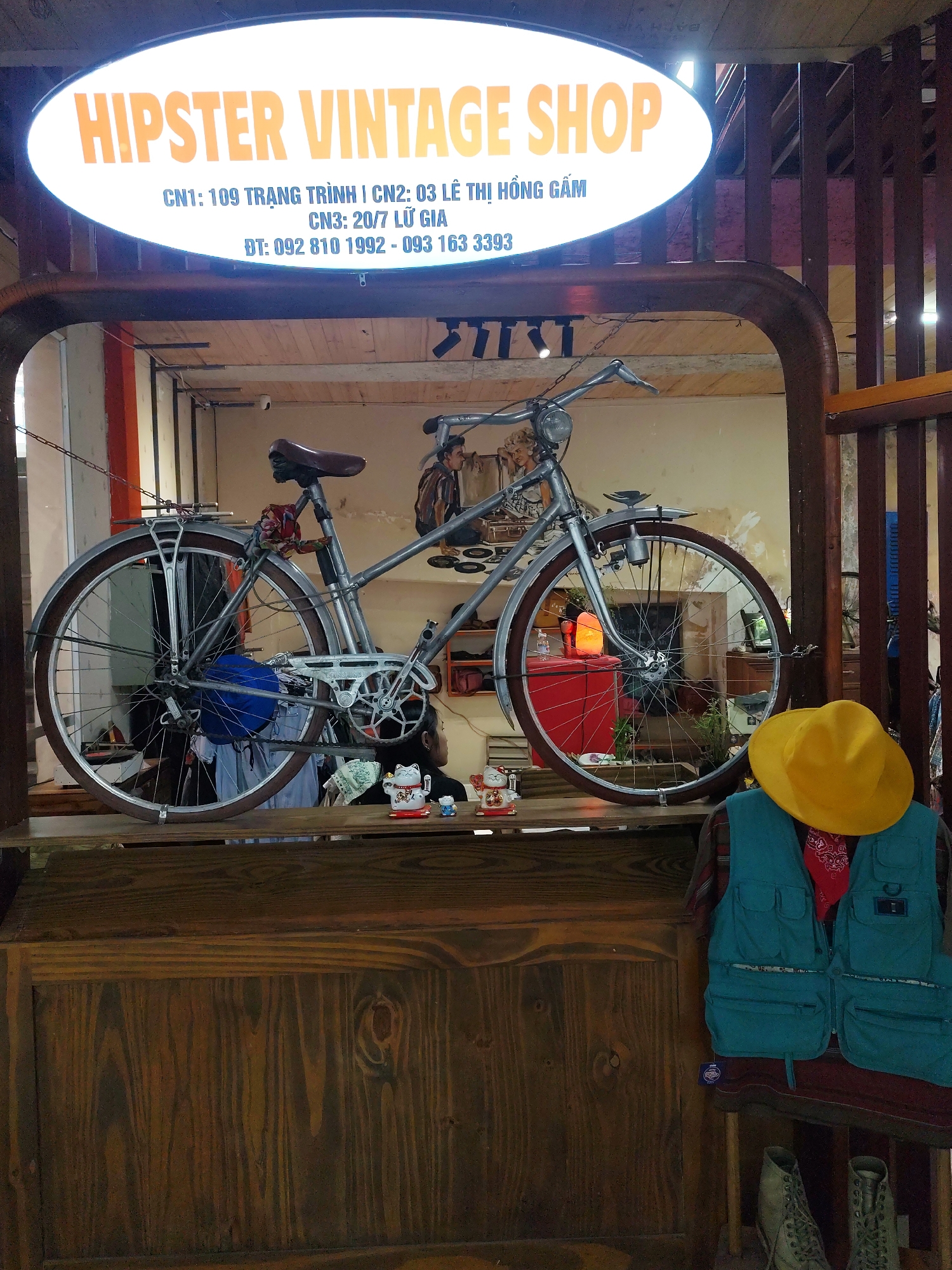 The image depicts a "Hipster Vintage Shop" display in Da Lat, Vietnam. A vintage bicycle is prominently displayed on a wooden shelf under an illuminated sign that reads "HIPSTER VINTAGE SHOP" along with three addresses and two phone numbers: CN1: 109 TRẠNG TRÌNH, CN2: 03 LÊ THỊ HỒNG GẤM, CN3: 20/7 LỮ GIA, and ĐT: 092 810 1992 - 093 163 3393. Below the bicycle, a mannequin wears a blue vest, a red bandana, and a yellow hat. A pair of boots sit at the base of the display. The shop's interior can be glimpsed behind the bicycle. The structure surrounding the display is made of dark wood.